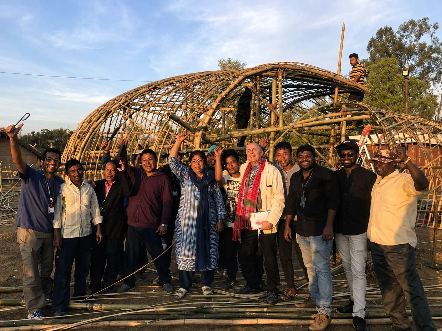 Group of people standing in front of a bamboo structure, celebrating and smiling during daylight.