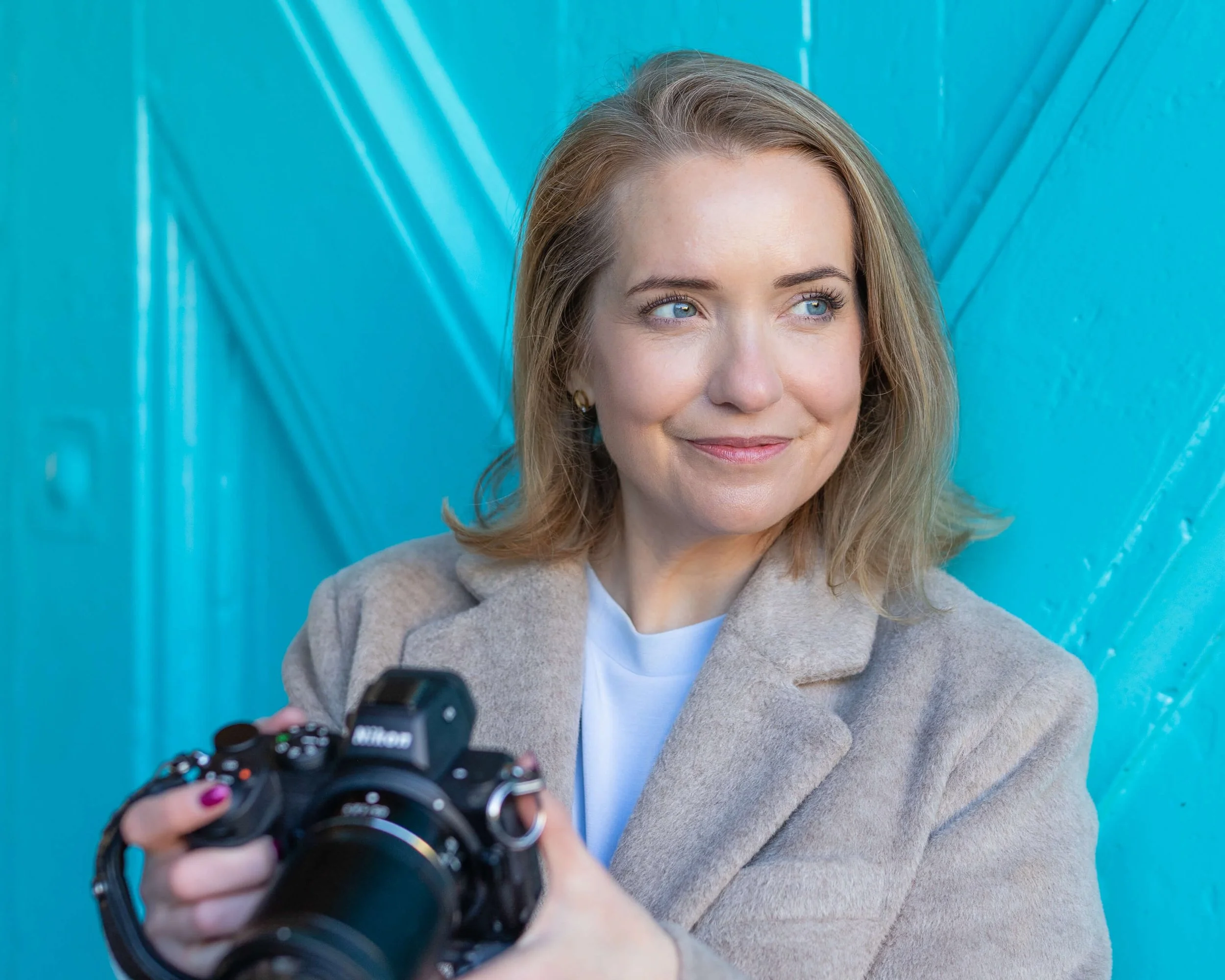A woman with shoulder-length blonde hair and blue eyes holding a Nikon camera, standing against a bright blue background.