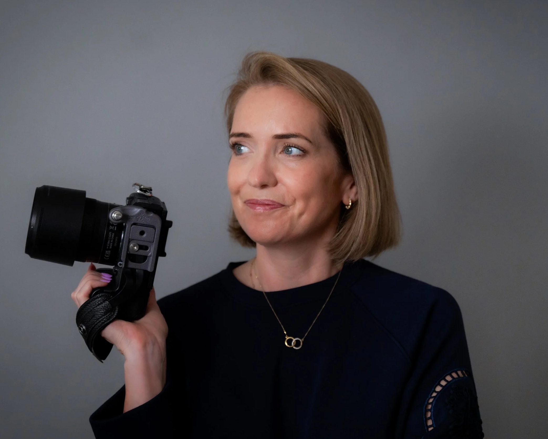 A woman with short blonde hair holding a camera in her right hand, wearing a black top with cutout details on the sleeve, gold earrings, and a necklace with two interlinked rings.