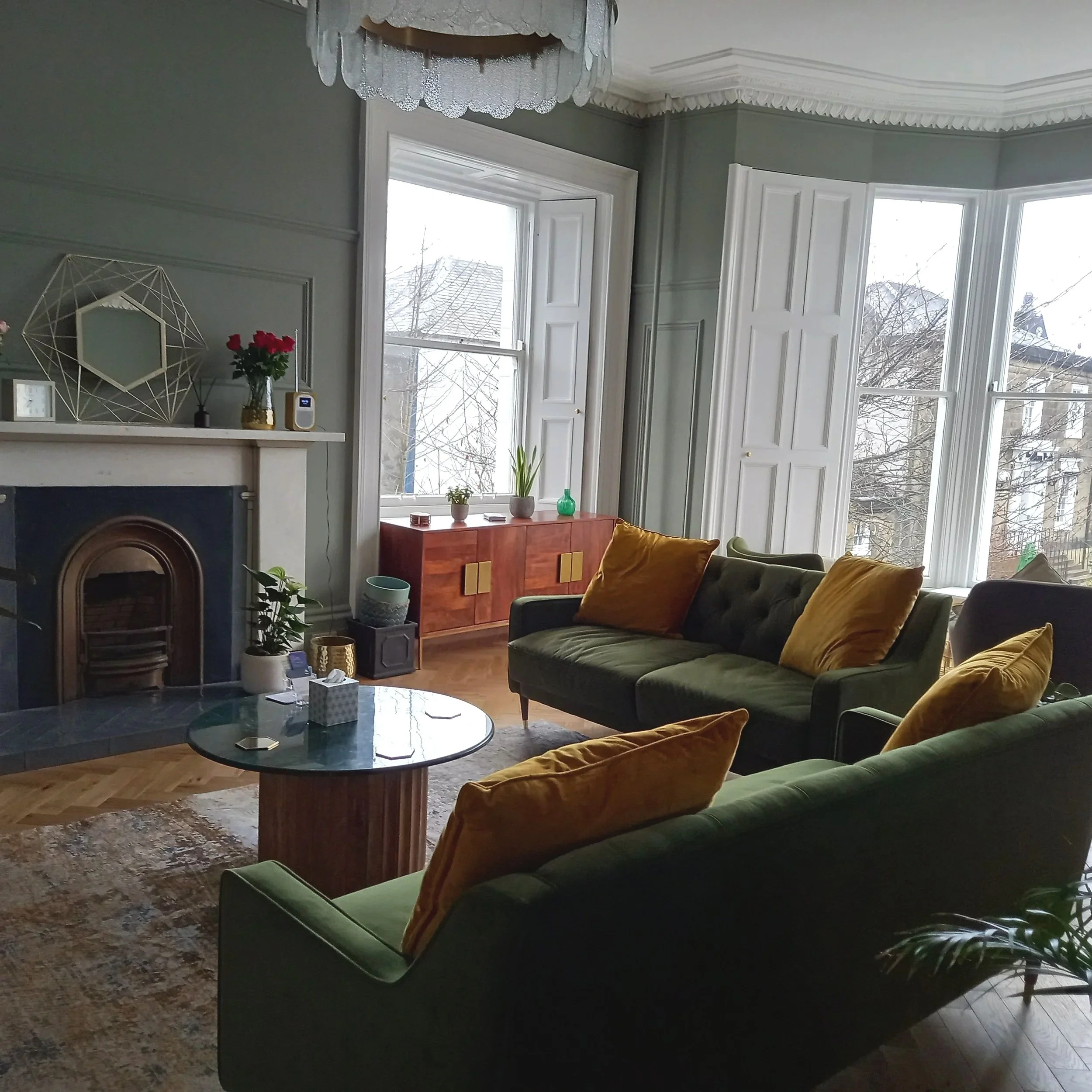 Two three seater leaf green settees with mustard yellow cushions in a L shape set back from a large bay window and near a fireplace. 