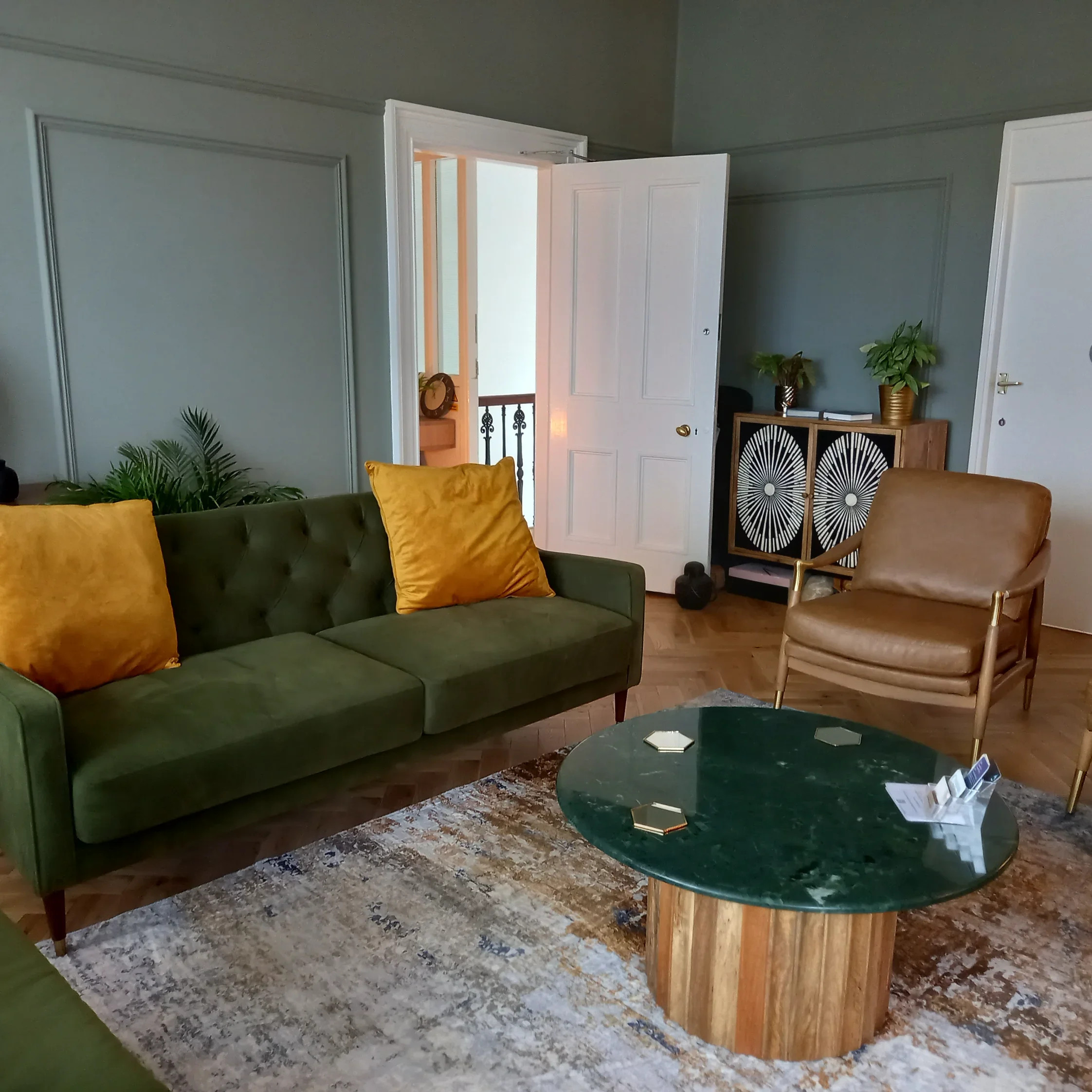 Waiting room with green sofa, brown armchair, marble-topped coffee table, and decorative cabinet against the wall. Open door reveals hallway with staircase. Plants and cushions are present.