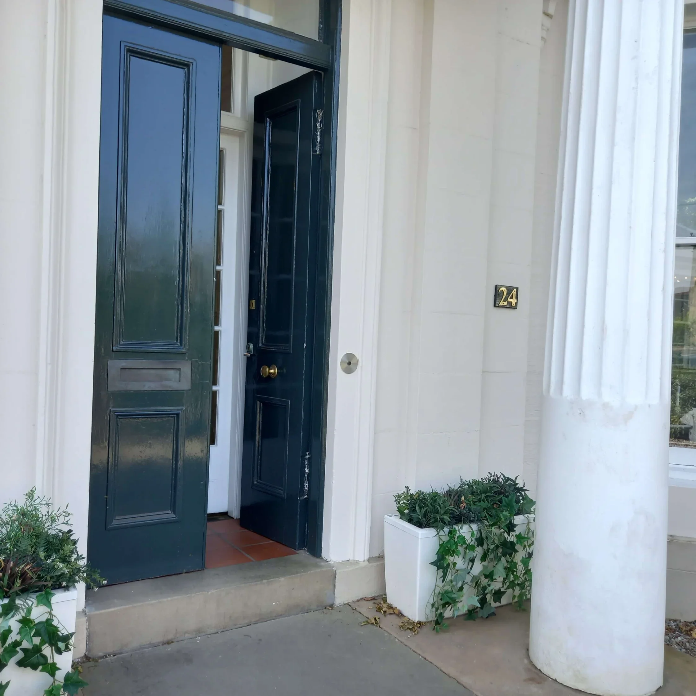 Open navy blue front door with gold doorknob, white house exterior with white column and house number 24, potted plants outside door.