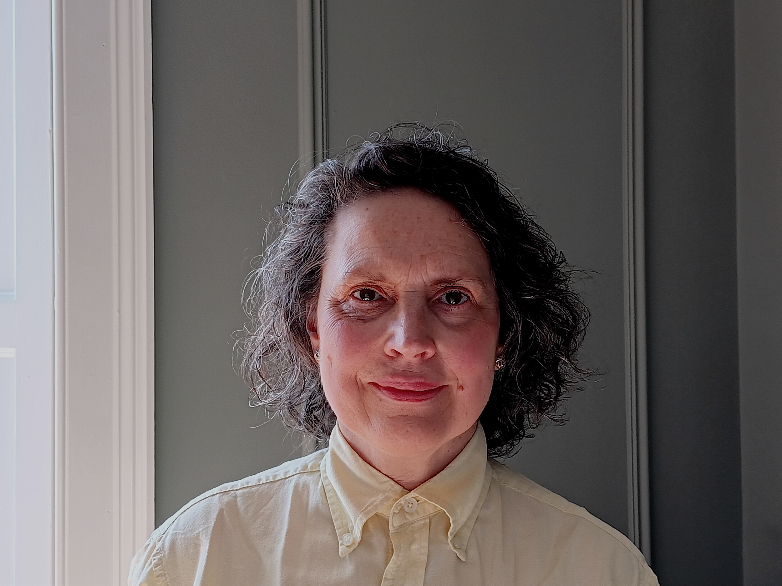 A woman with curly hair is wearing a butter yellow collared shirt. She is standing indoors in front of wood panelling painted grey, with a white window visible on the left side of the image.