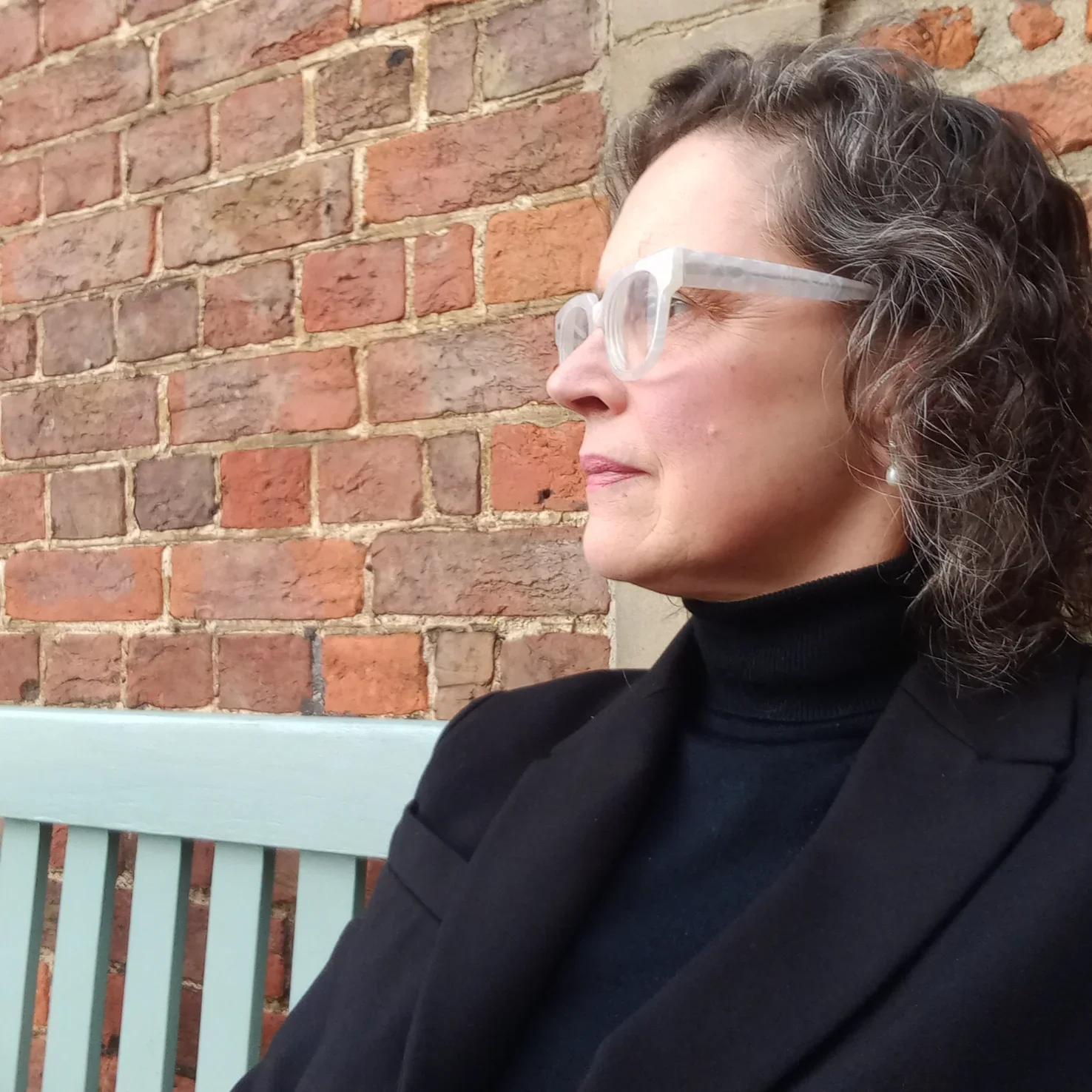 A woman with curly brown hair and glasses with clear frames, wearing a black turtleneck and black blazer, sitting on a light-colored bench against a brick wall, looking to her left.