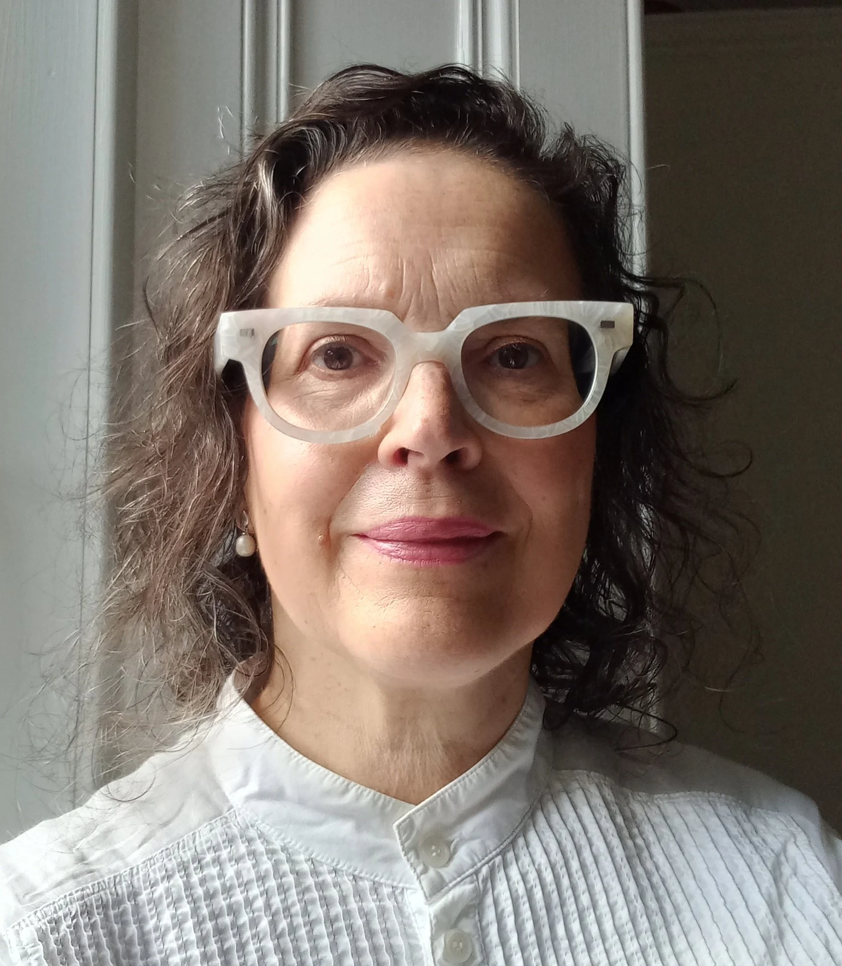 A woman with curly brown hair wearing large white glasses and a white blouse with vertical pleats.