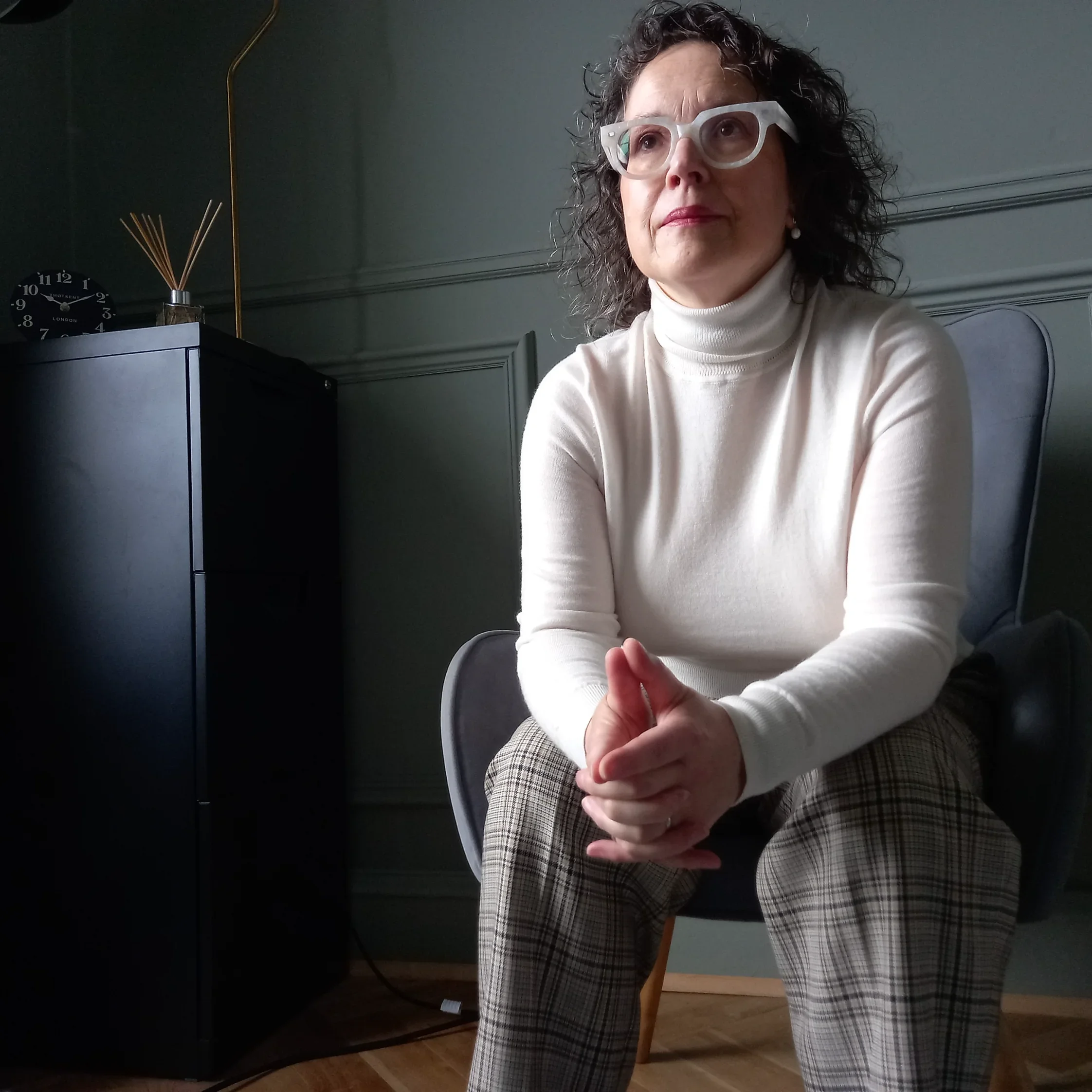 A woman with curly hair wearing large clear glasses, a white turtleneck, and plaid pants, sitting on a chair in a room with dark walls and a wooden floor, with a black cabinet, clock, and reed diffuser in the background.