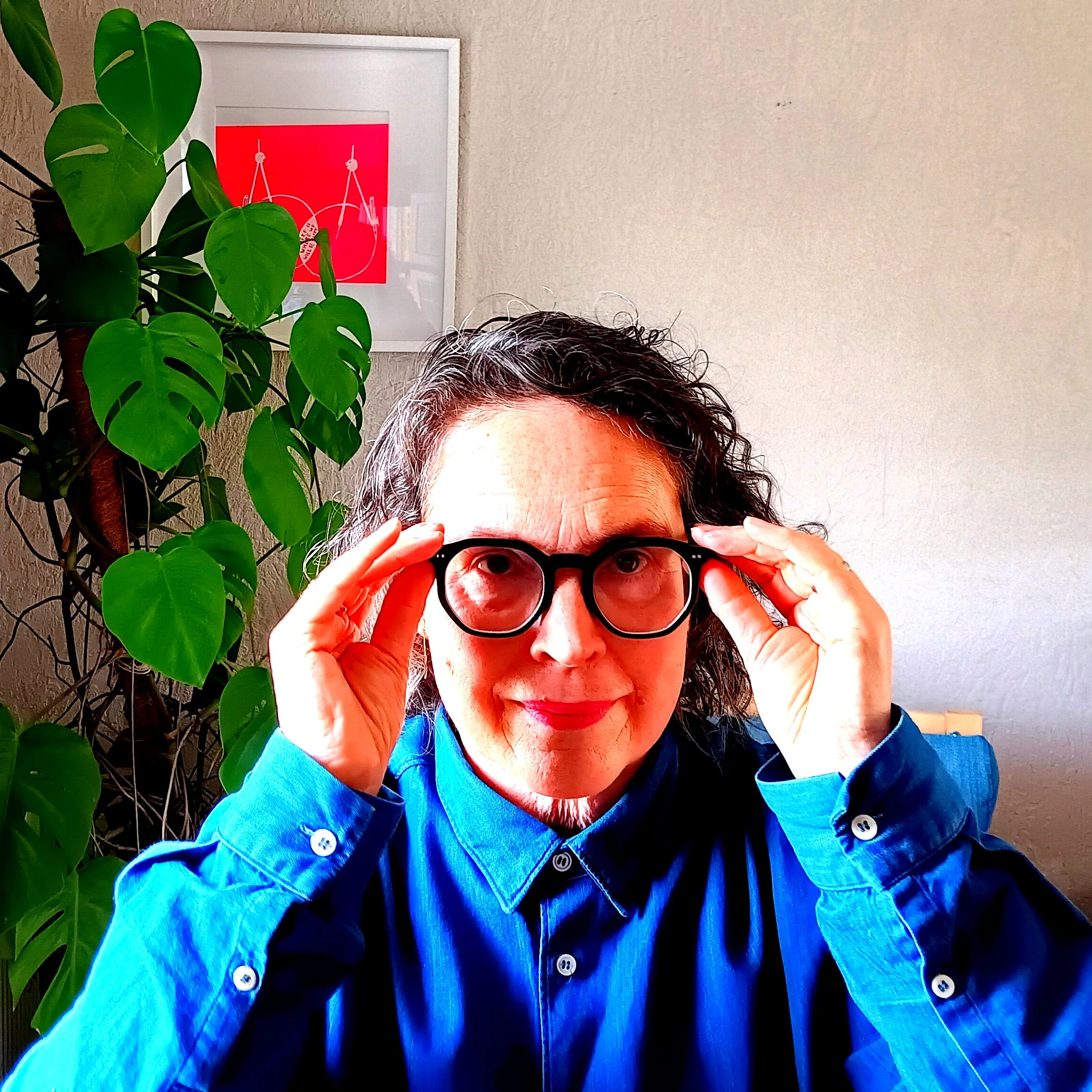 Person with curly dark hair and a blue button-down shirt, putting on reading glasses, sitting in front of a tall Monstera plant and a  Rob Ryan 'compassion' print.