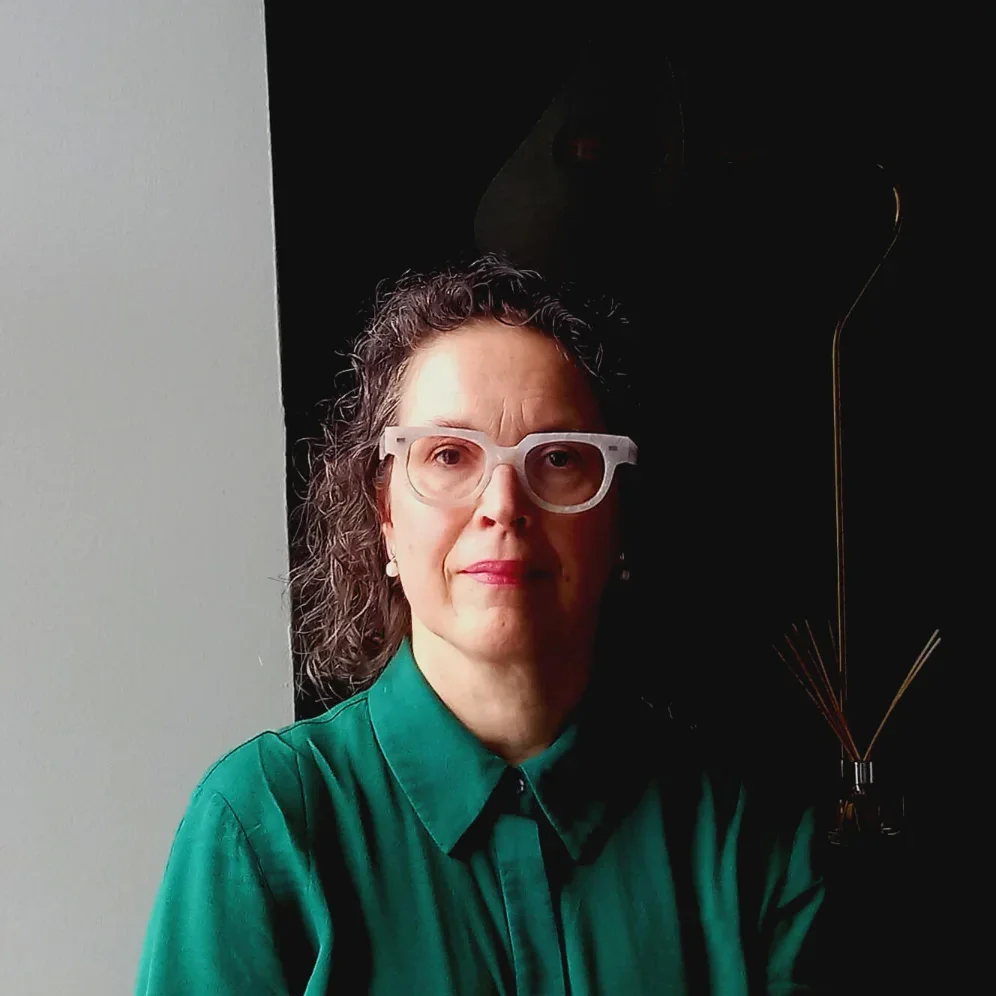 A woman with curly brown hair and clear glasses wearing a green button-up shirt, standing indoors against a darker background.