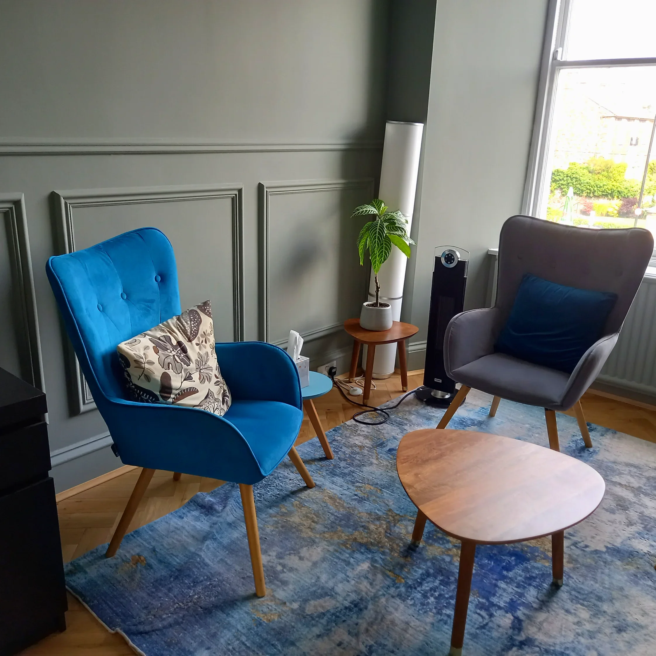 A cosy therapy room corner with two modern chairs, one blue with a patterned cushion and one grey with a dark blue pillow, a small wooden coffee table, a potted plant, and a window with a view of trees and outside architecture.