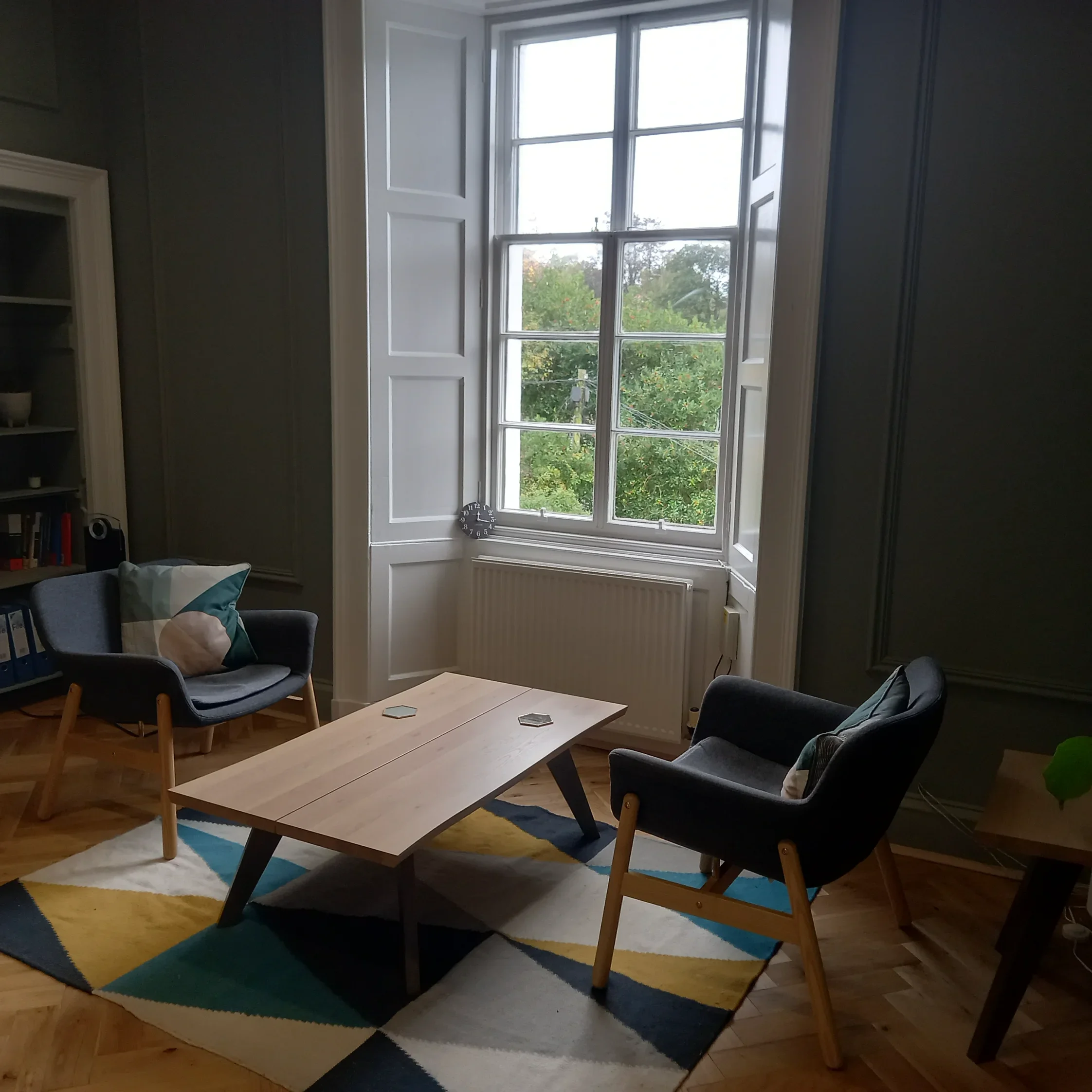 A naturally lit therapy room with a wooden coffee table, two dark armchairs with patterned pillows, a colourful geometric rug, large window with white framing, and a small bookshelf on the side.