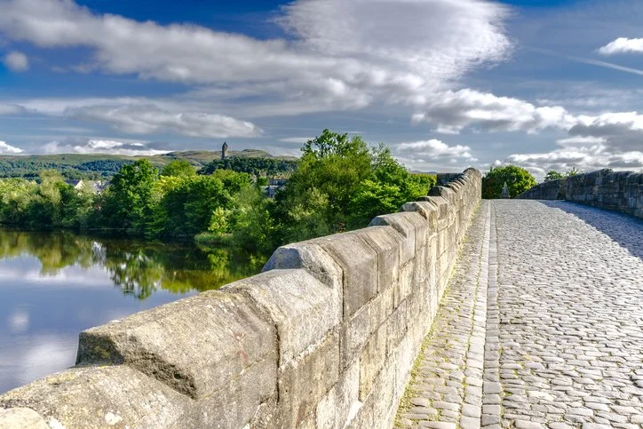 Stone bridge over a river with lush green trees, a cloudy sky, and distant hills.