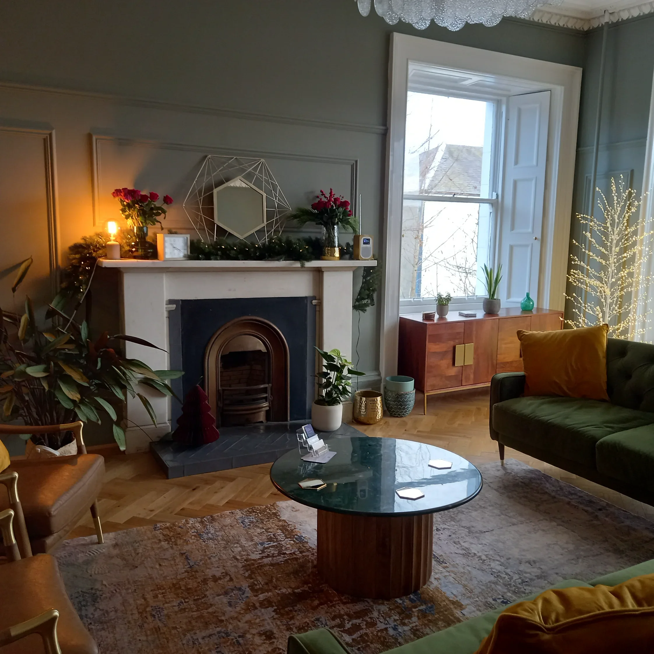 There are plants near the fireplace and a glass-topped coffee table in the centre of the waiting room. A large window with blinds is on the right, and a wooden sideboard with small potted plants is beneath it. 