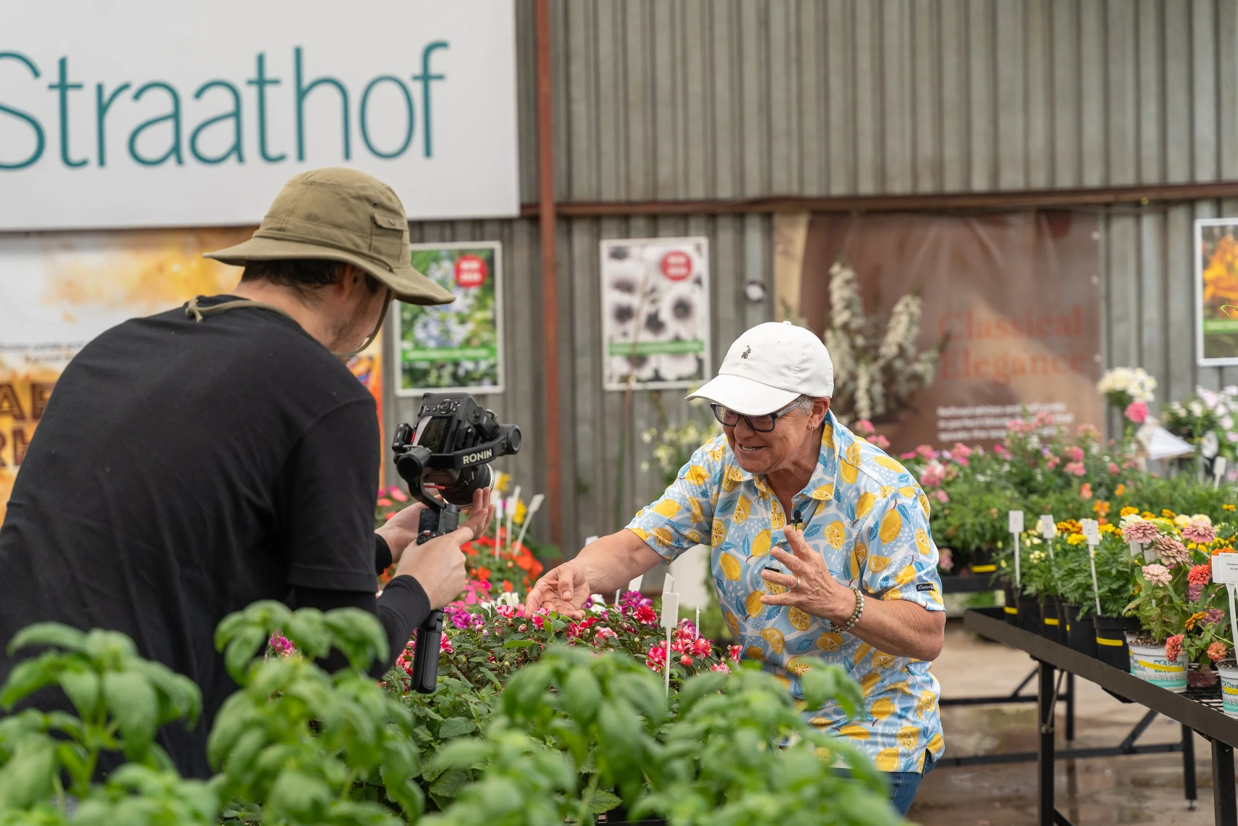 Tanya Visser in a lemon-patterned shirt and white hat showing a flower to Gareth filming with a camera in a greenhouse filled with colorful flowers.