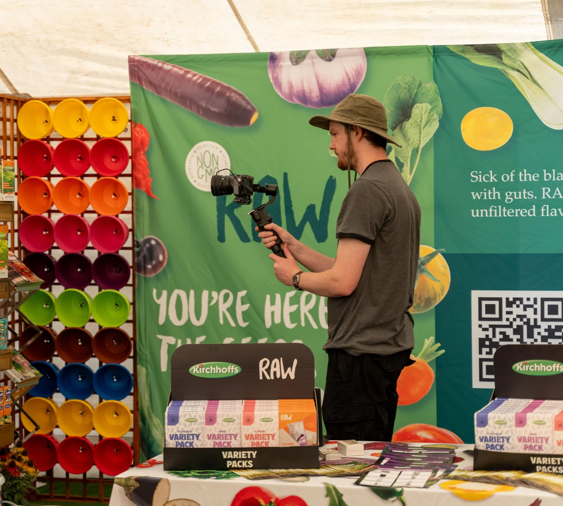 A man filming with a camera stabilizer at an herbal or natural foods market booth with a green banner behind him that reads "YOU'RE HERE" and features vegetables and a QR code. Colorful buckets are on a display to his left, and various packaged herbal packs are on the table in front of him.