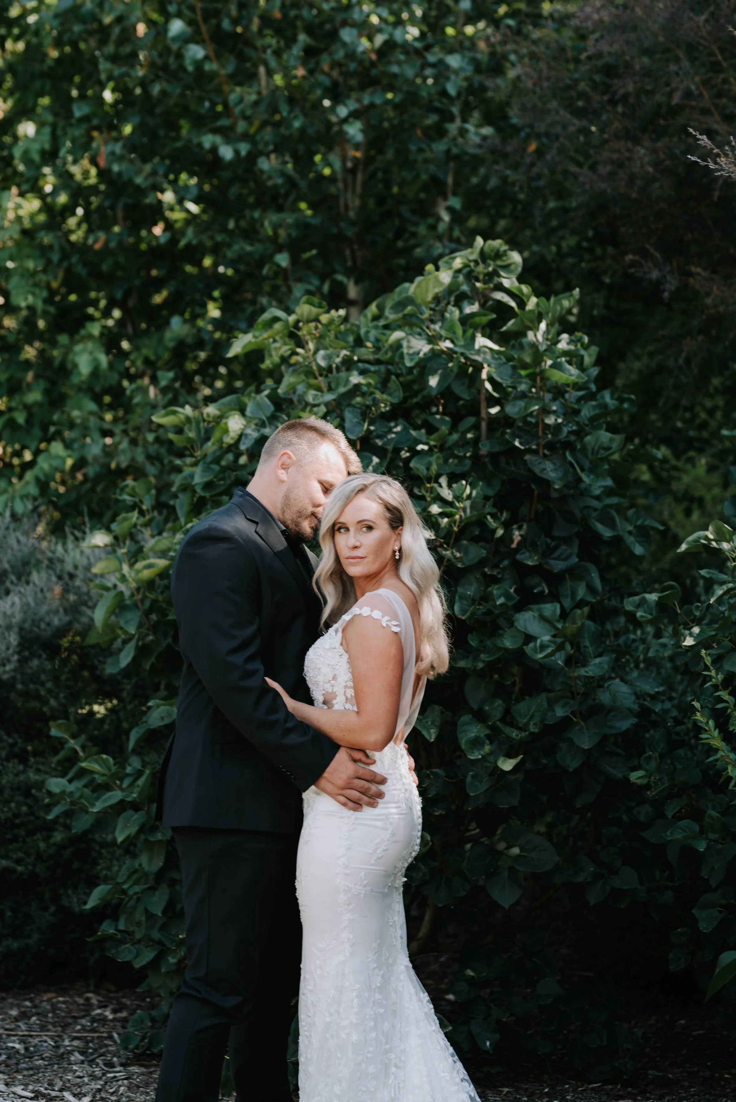 Bride and groom standing close together outdoors, surrounded by lush greenery, in a romantic pose.