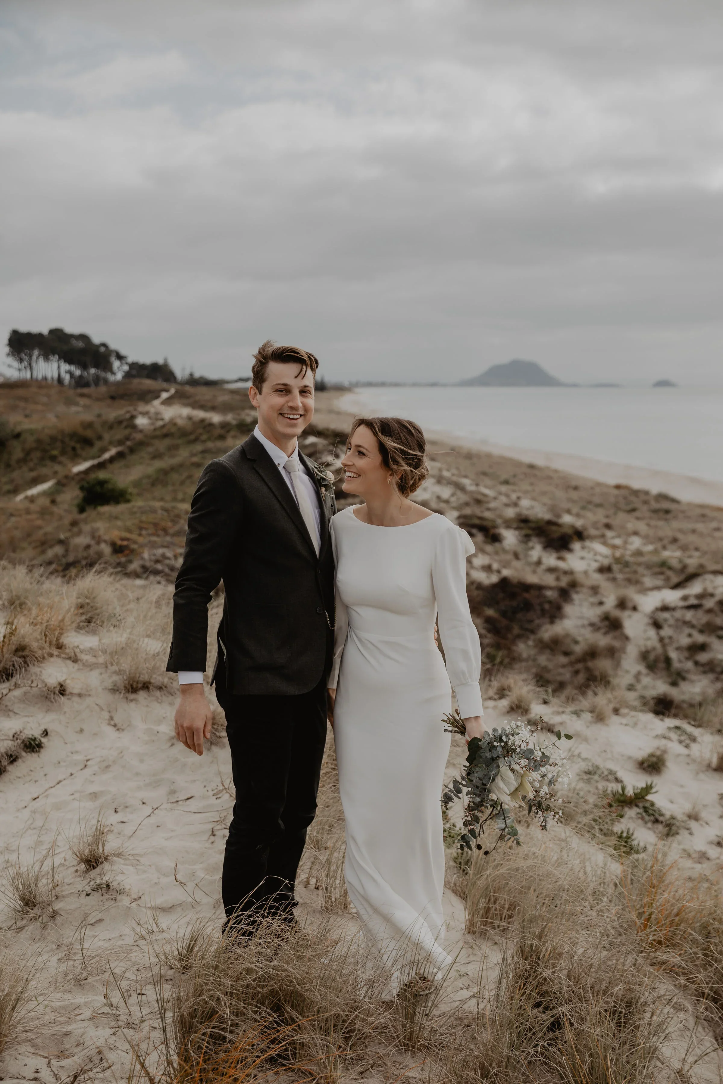 A newlywed couple in wedding attire standing on a sandy beach with grass and dunes, holding a bouquet, smiling, with ocean and cloudy sky in the background.