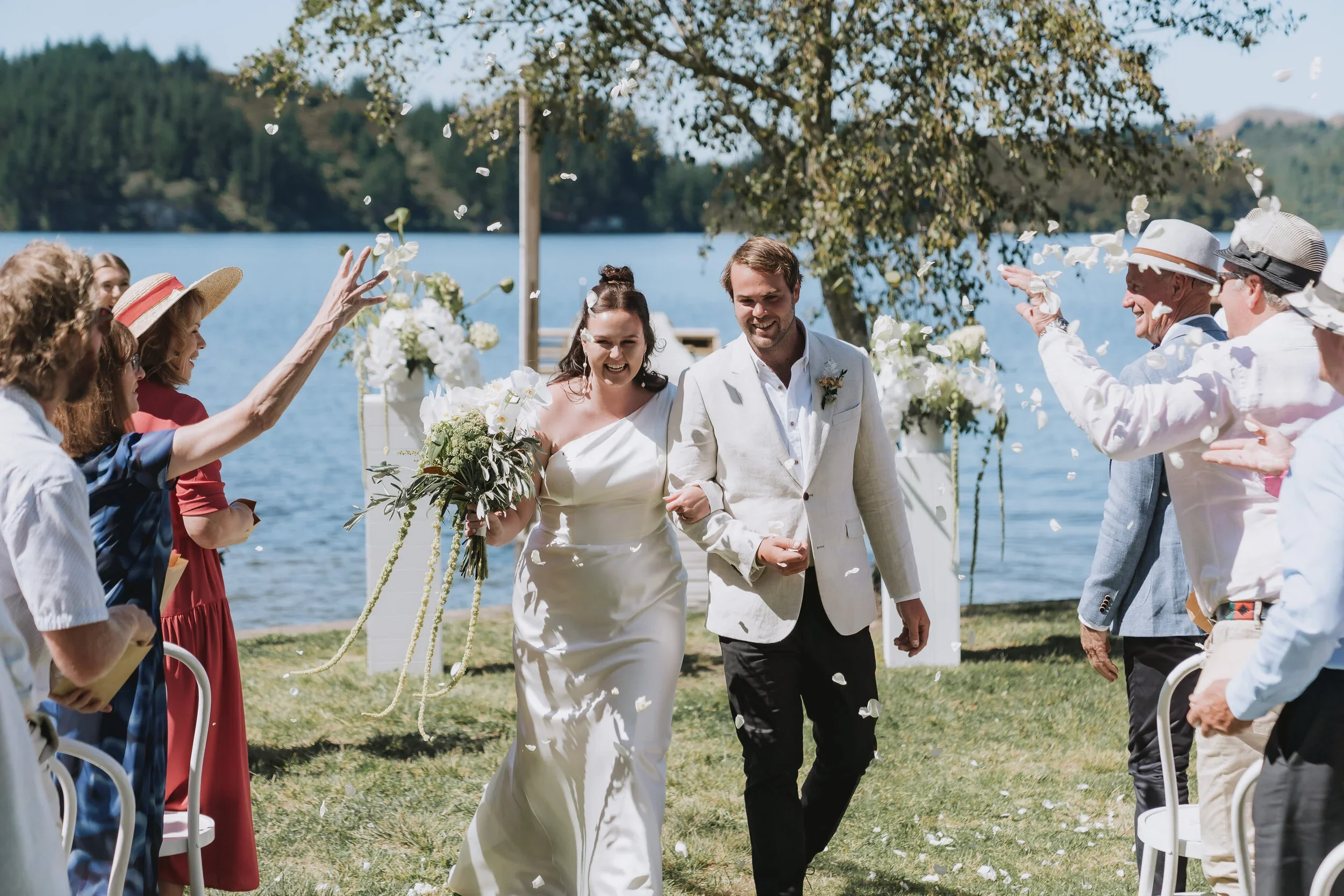 Bride and groom walking down aisle outdoors by the lake, celebrating wedding with friends, flower petals being tossed