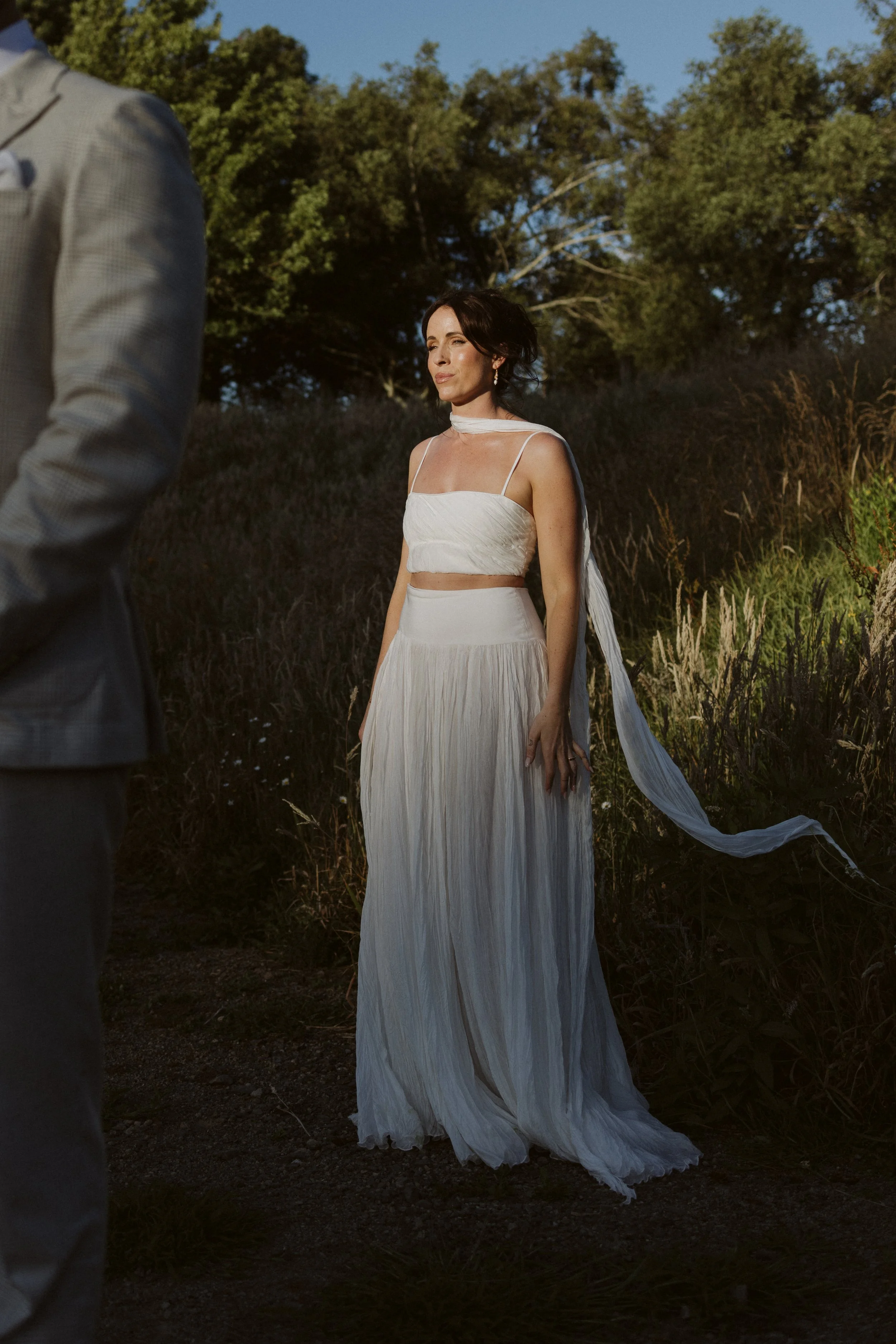 A woman in a white dress standing outdoors in a field with trees in the background, illuminated by sunlight.