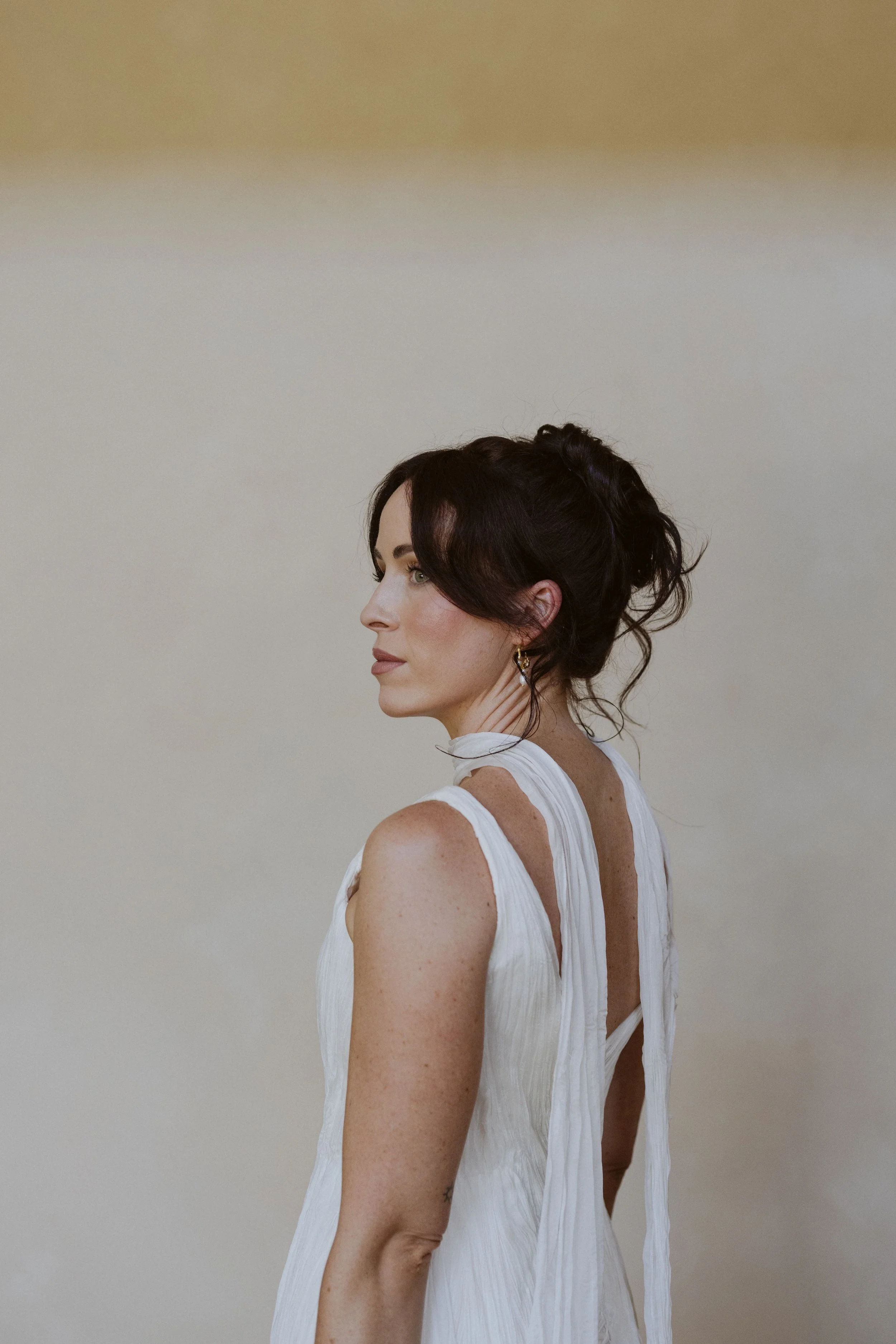 A woman with dark, wavy hair styled in an updo, wearing a sleeveless white dress with pleats and a tie at the back, standing sideways against a plain neutral background.