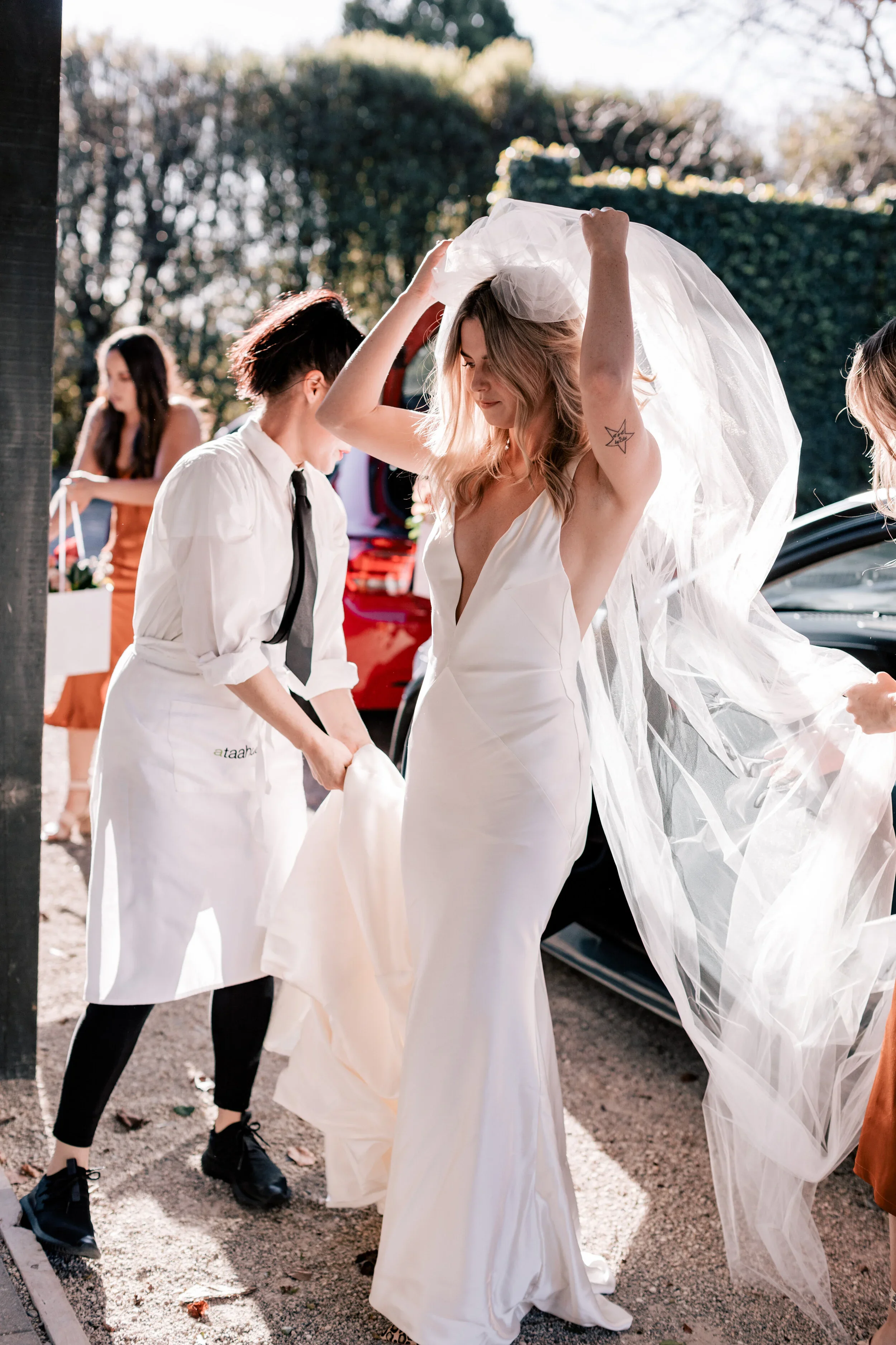 A woman in a white wedding dress is putting on her veil outside, with assistance from a staff member. Other women and a red car are visible in the background.