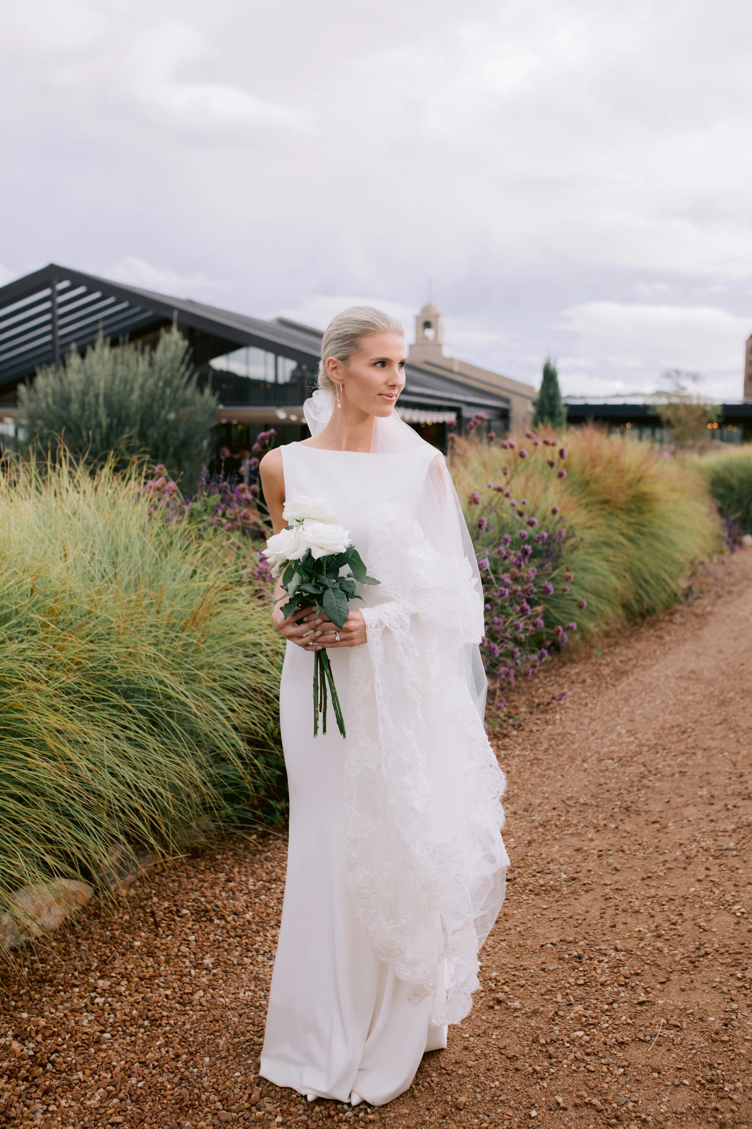 A bride in a white wedding dress holding a bouquet of white roses stands on a dirt path surrounded by tall green and purple plants, with a modern building and cloudy sky in the background.