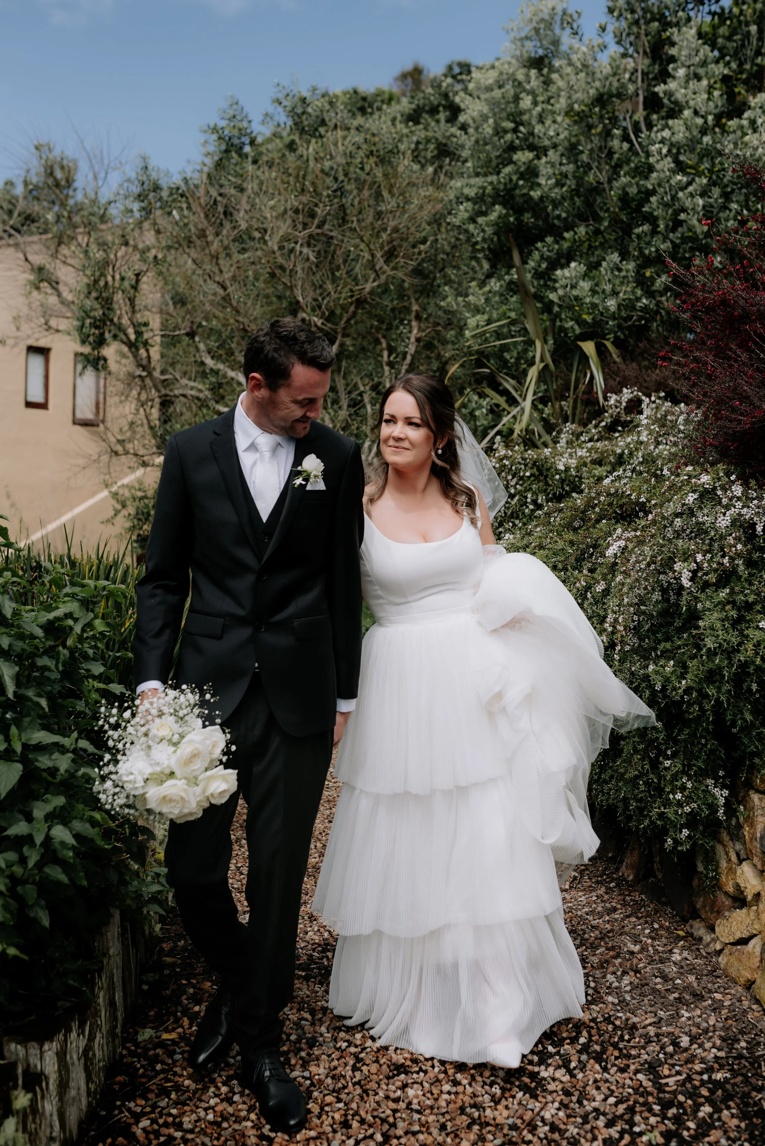 A bride and groom walking outdoors on a gravel path surrounded by greenery and flowering bushes, with the groom holding a bouquet of white roses.
