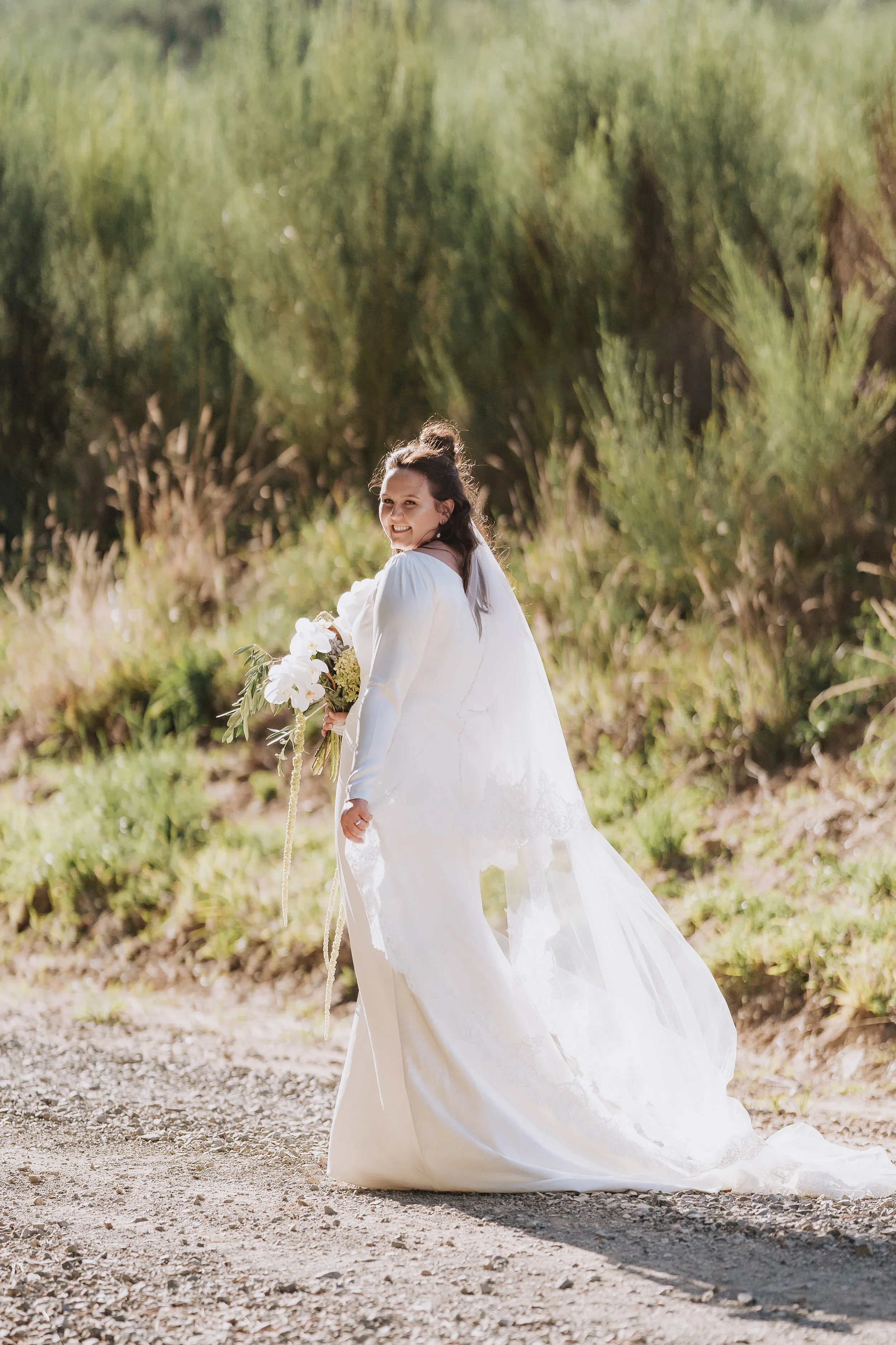 A woman in a wedding dress walking outdoors in a natural setting with greenery, holding a bouquet of flowers, smiling at the camera.