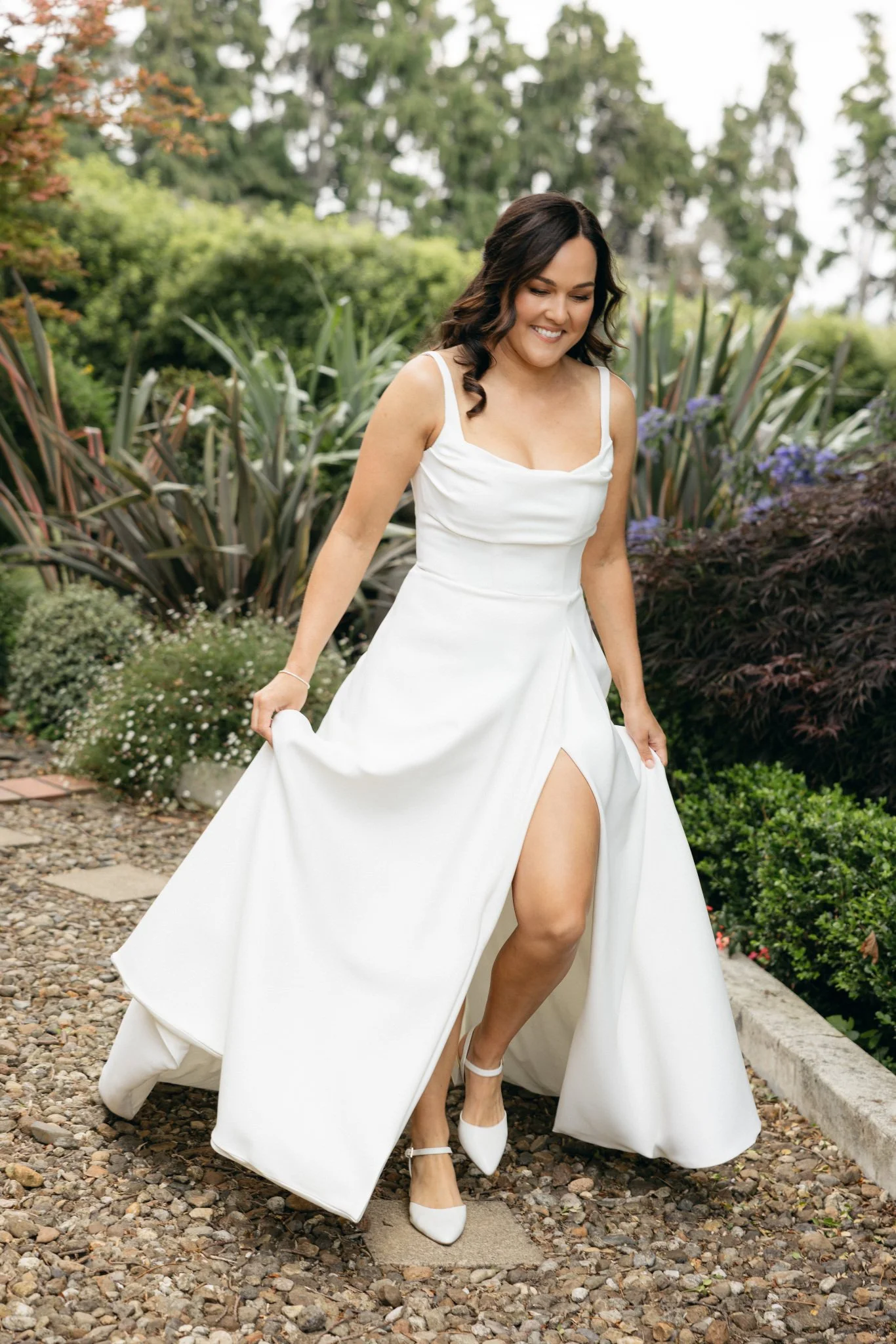 A woman in a white dress and white heels outdoors, lifting her dress to show her leg and smiling. She has dark hair styled in loose waves, surrounded by lush green plants and trees.