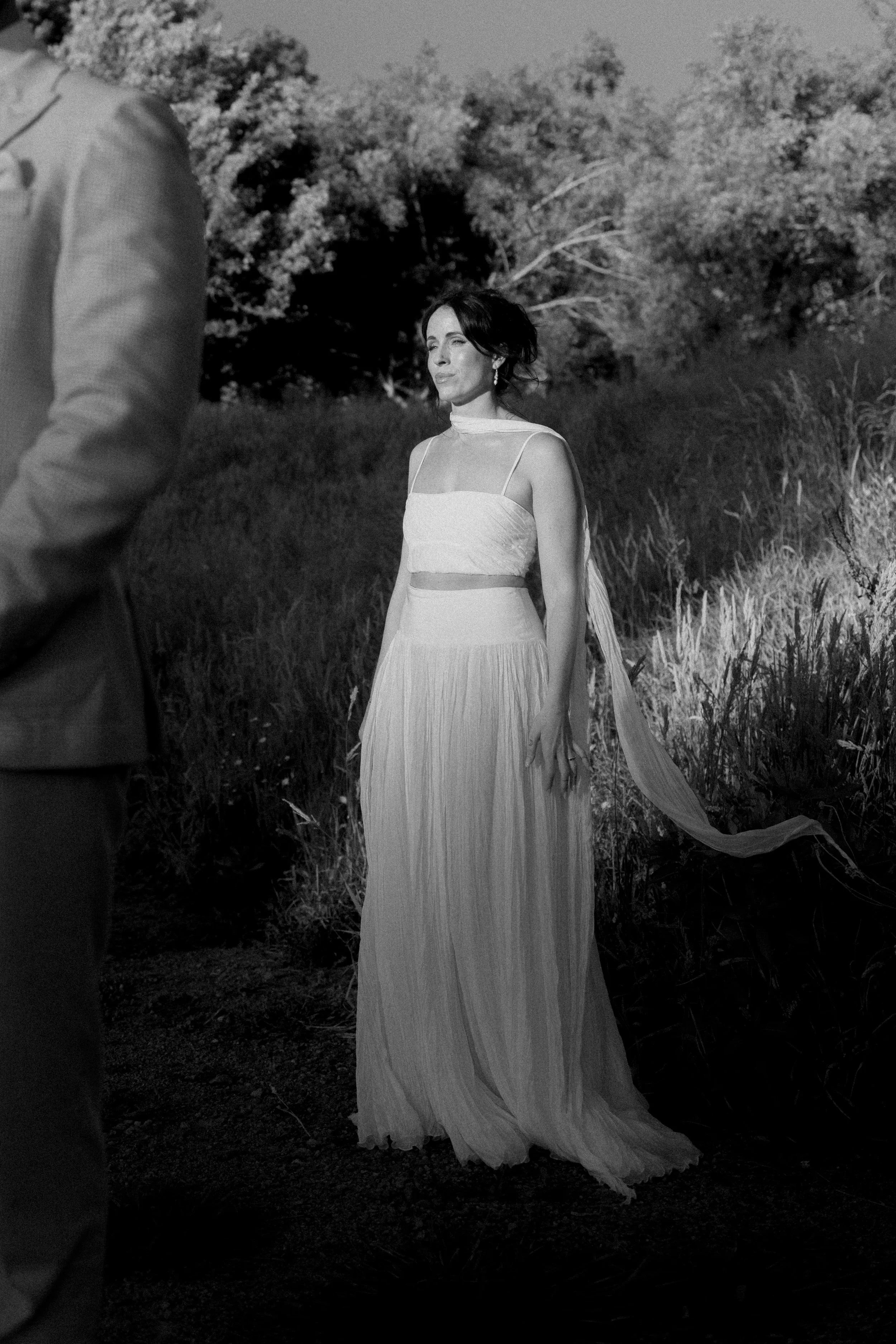 A woman in a long, flowing dress stands outdoors near tall grass and trees, with an overcast sky behind her.