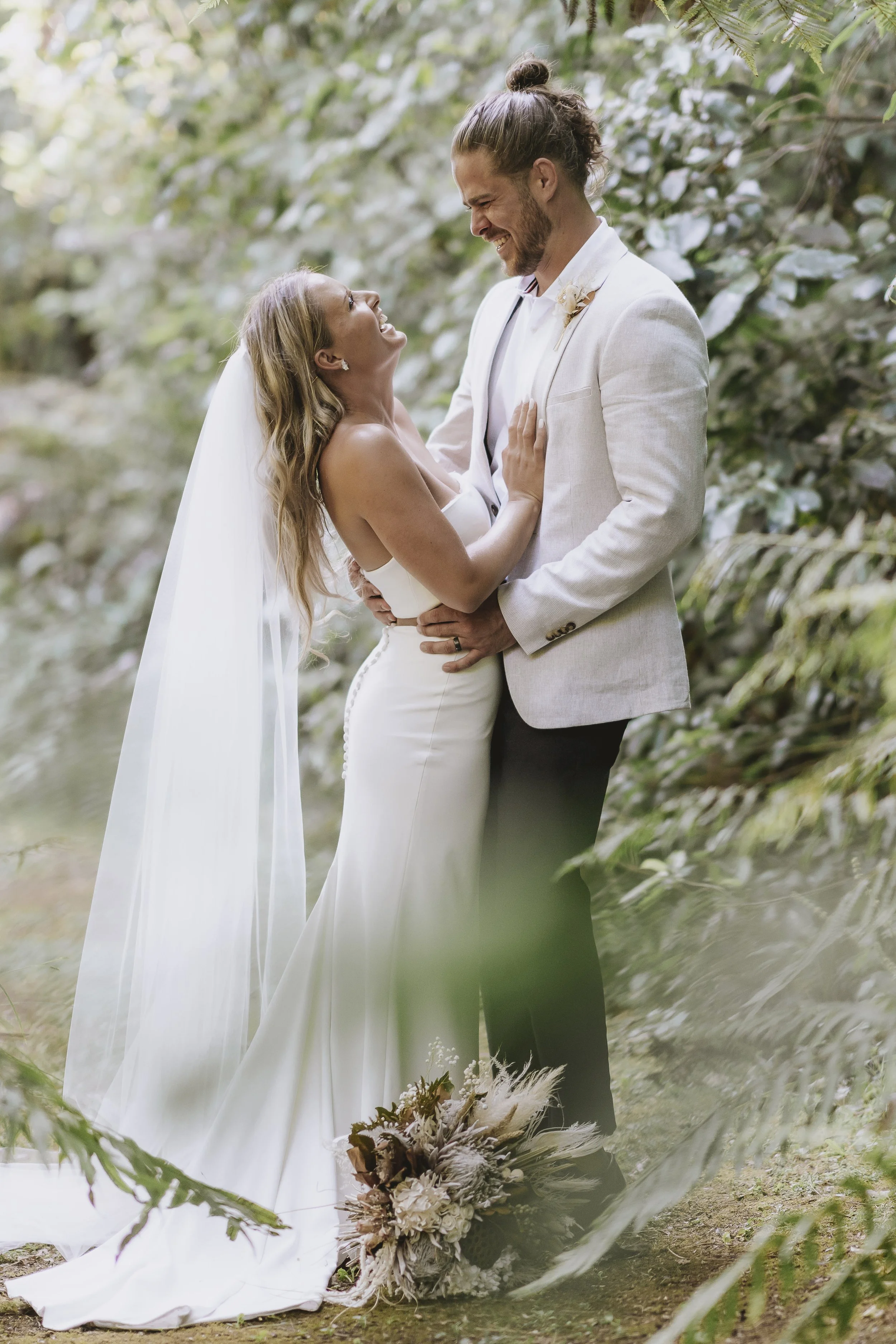 A bride and groom share a joyful moment outdoors, surrounded by greenery, with the bride wearing a white gown and veil, and the groom in a light-colored suit with a flower boutonniere.
