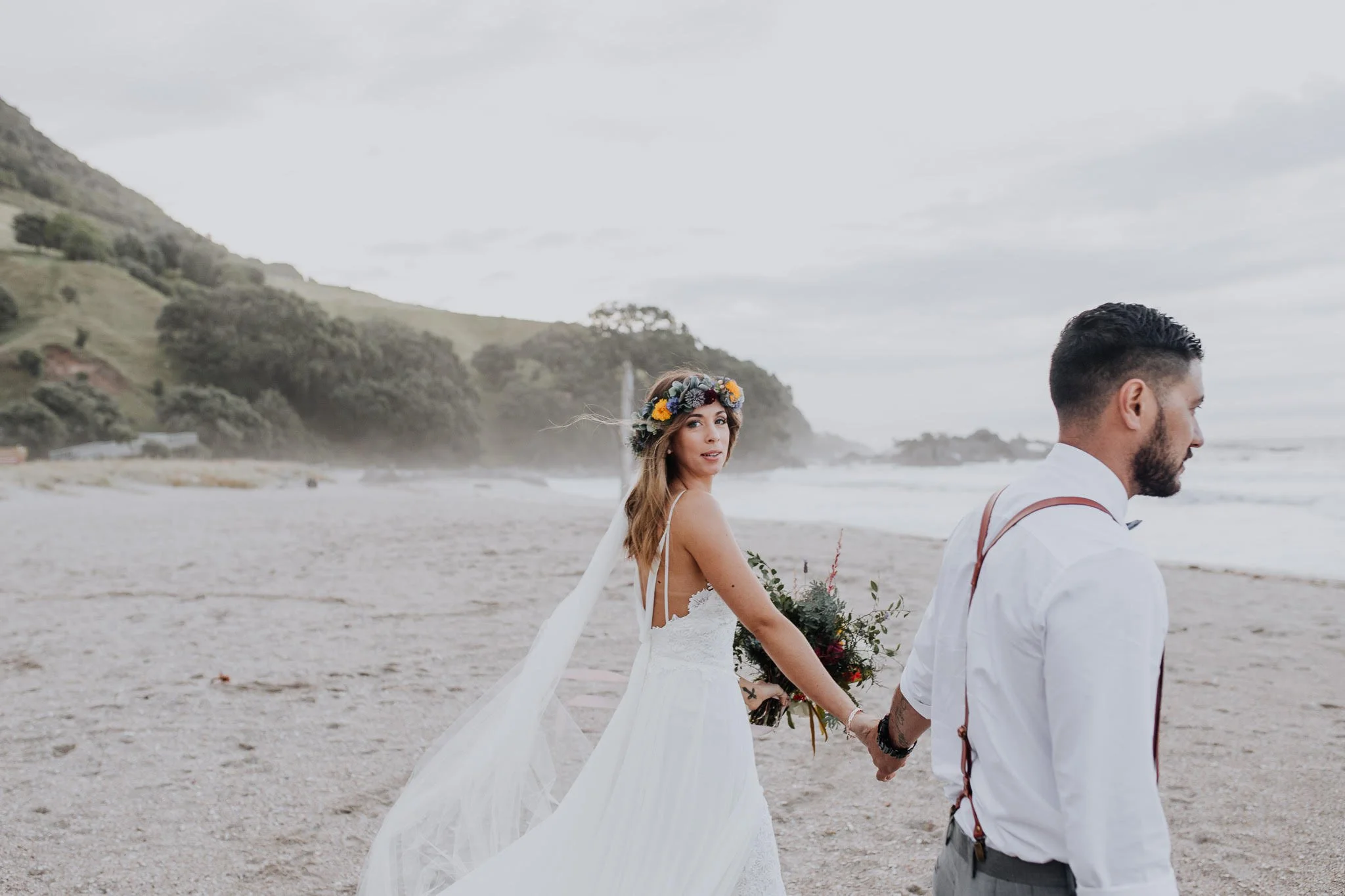 A bride and groom holding hands on a beach; the bride wears a white dress and floral crown, the groom wears a white shirt with suspenders, with mountains and ocean in the background.