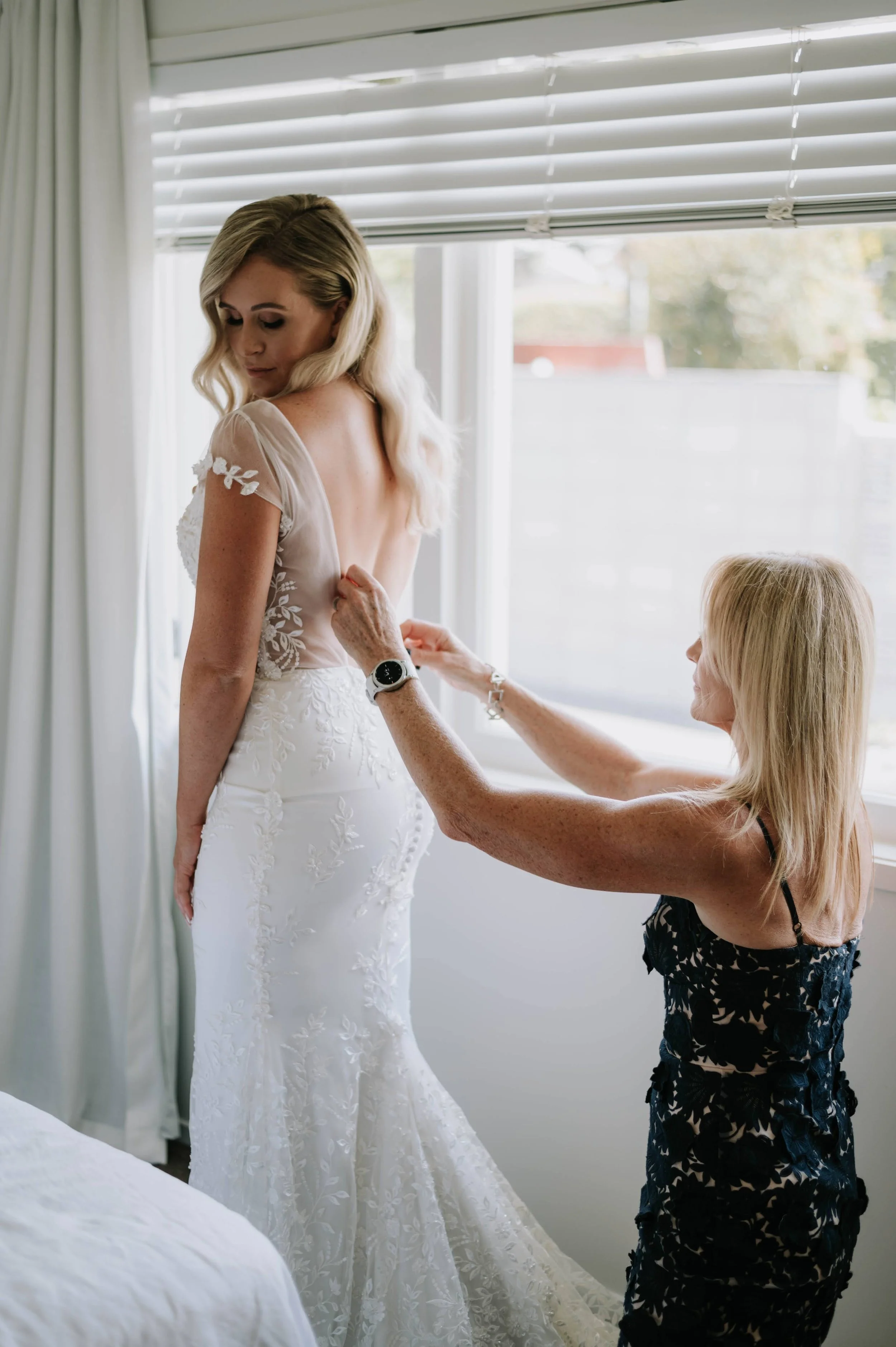 A bride in a white lace wedding dress is being helped into her gown by an older woman near a window with white blinds.