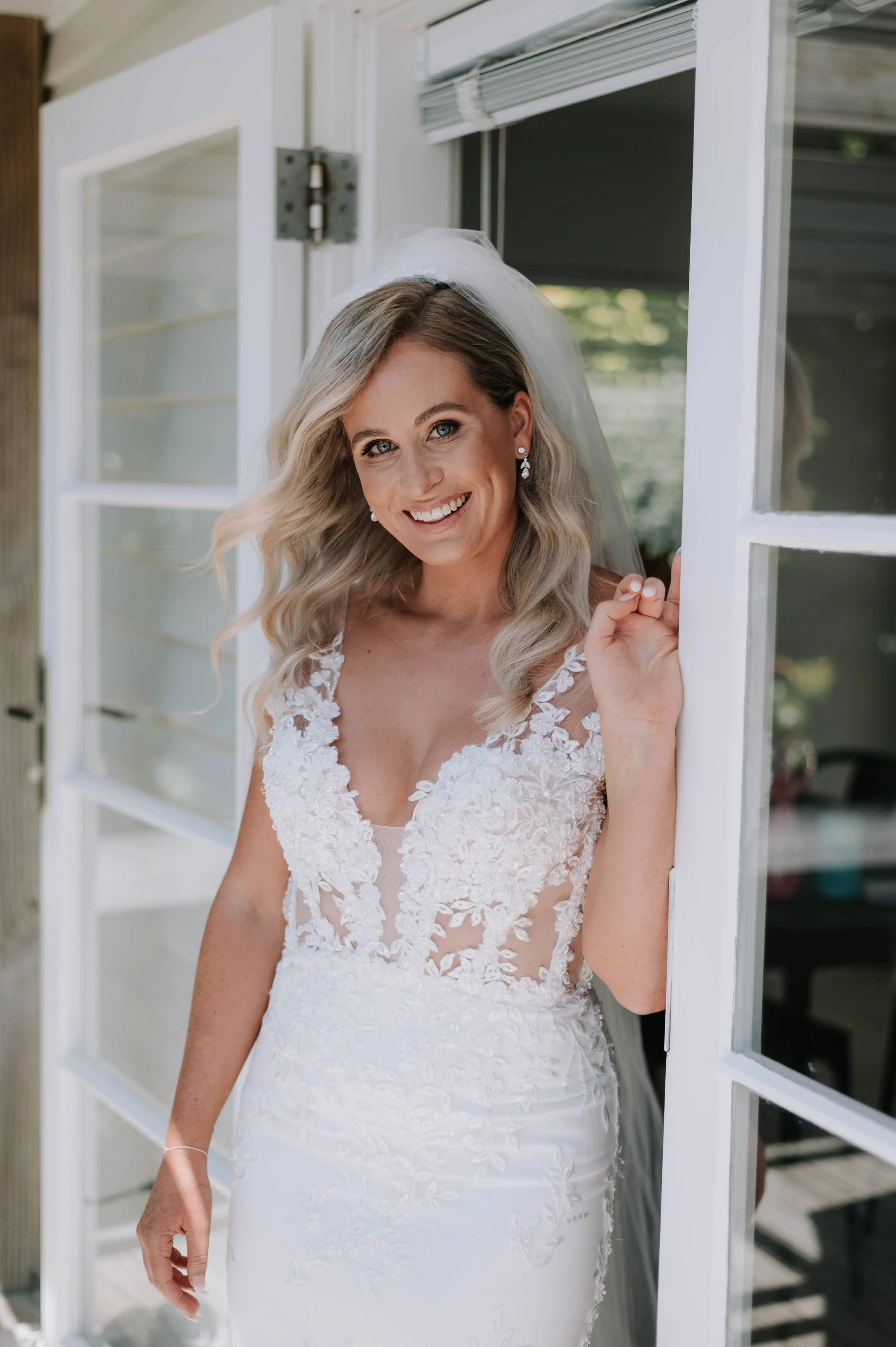 A smiling woman in a wedding dress standing by an open door, with a veil and earrings.
