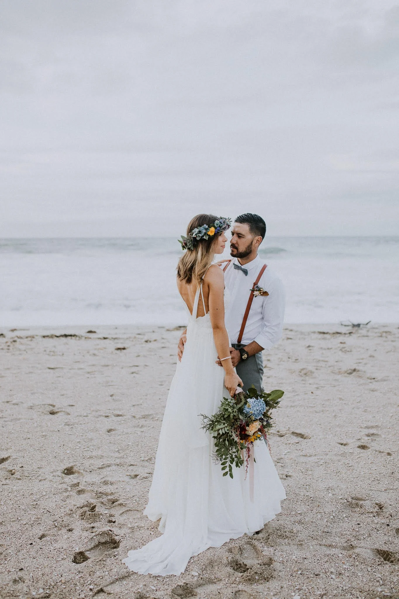 A couple dressed in wedding attire standing on a beach, facing each other with the ocean in the background. The bride is holding a bouquet of flowers and wearing a flower crown, while the groom is dressed in a shirt with suspenders and a bow tie.