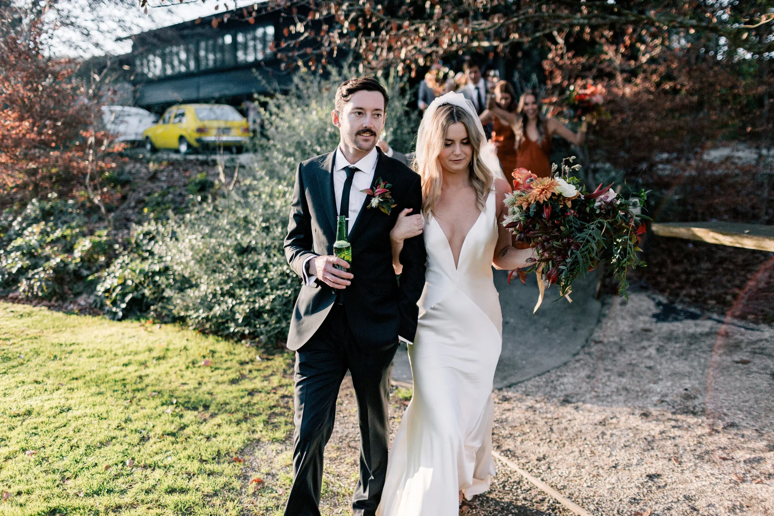Bride and groom walking together outdoors after wedding, with bridesmaids in the background, holding a bouquet of flowers, and a pickup truck and yellow car parked on a hill.