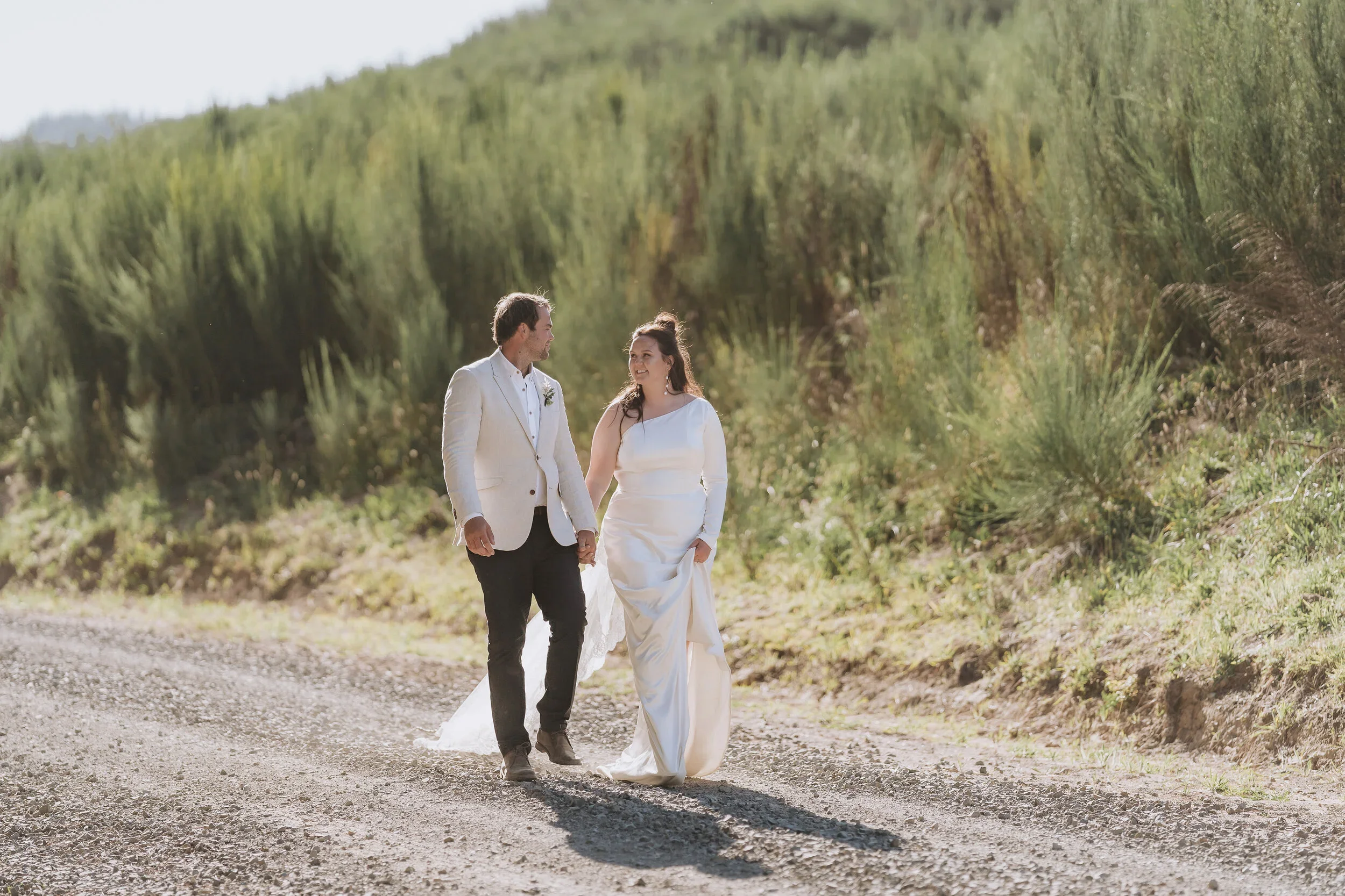 A bride and groom walking hand in hand on a dirt road, surrounded by green hills and bushes, on a sunny day.