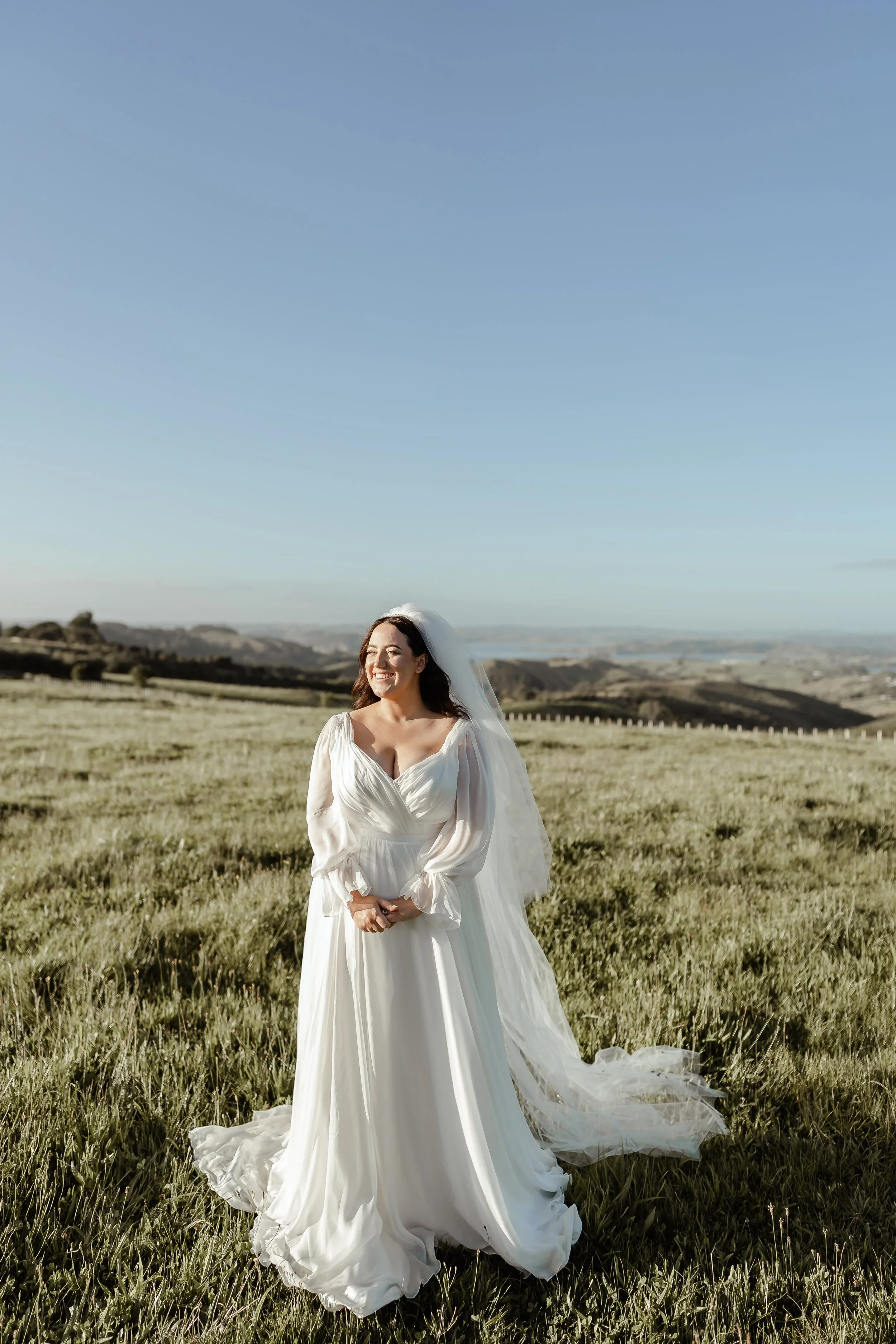 A woman in a white wedding dress standing in a grassy field with rolling hills in the background and a clear blue sky.