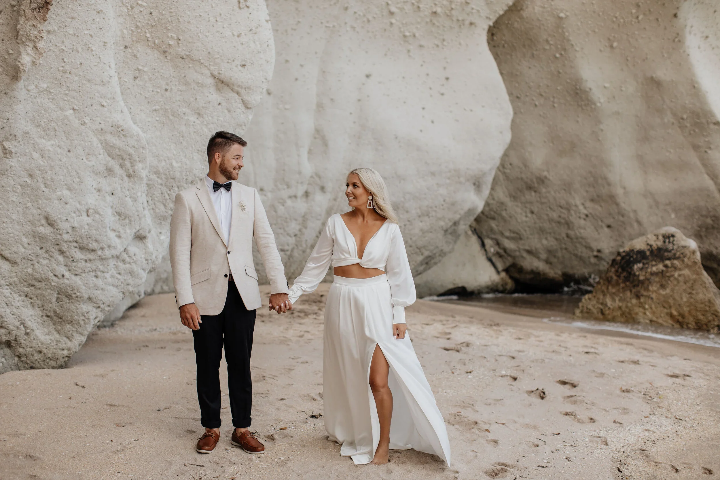 A couple in wedding attire holding hands on a sandy beach with large rocks and cliffs in the background.