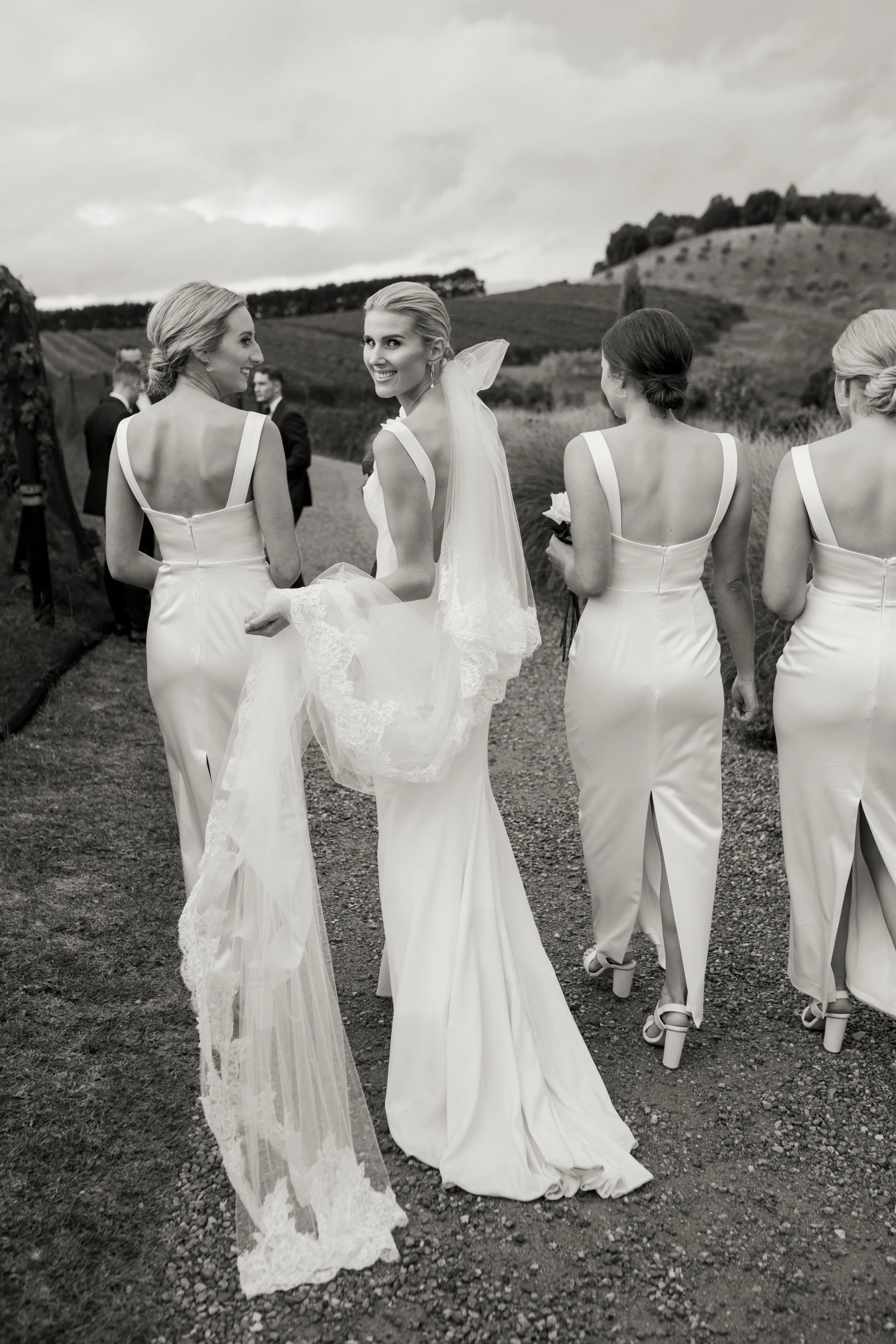 A bride and her bridesmaids walk outdoors in a vineyard, wearing white dresses. The bride is in the center, smiling and holding her veil, as the other women walk behind her with their backs turned.