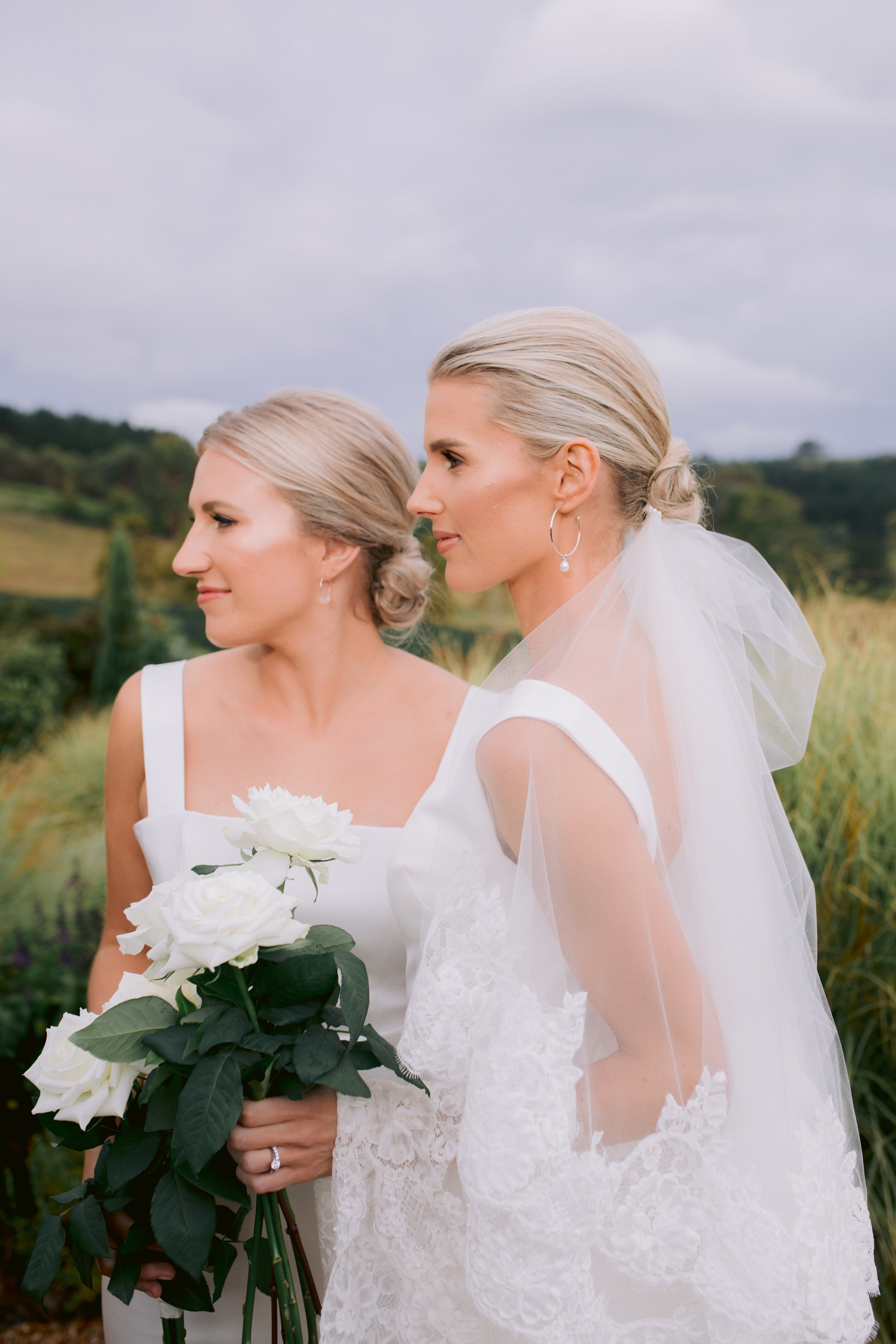 Two women in white wedding dresses, one holding a bouquet of white roses, standing outdoors in a field with greenery and hills in the background.