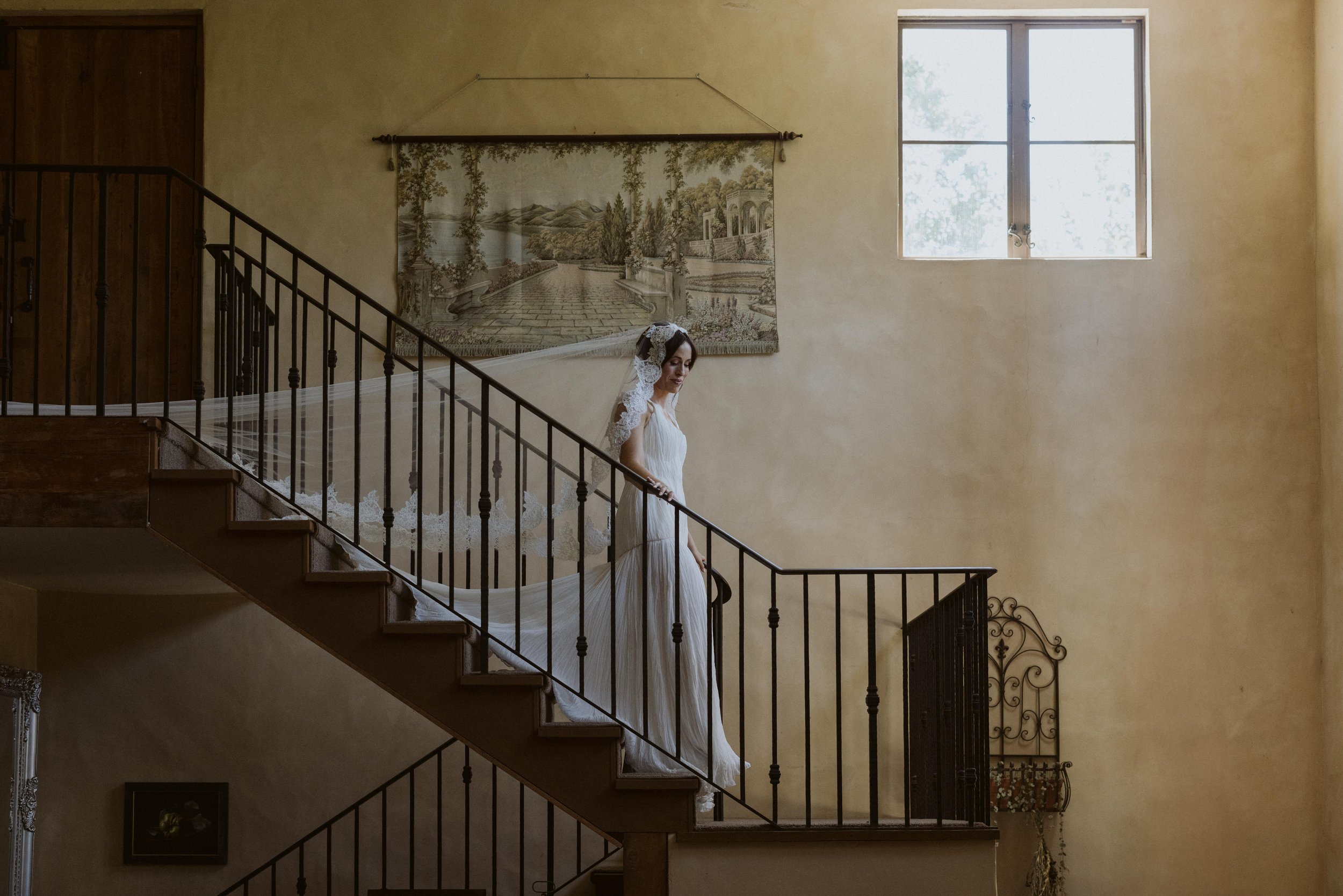 A bride in a white wedding dress and veil walking down a staircase inside a home with yellow walls and a window that lets in natural light.