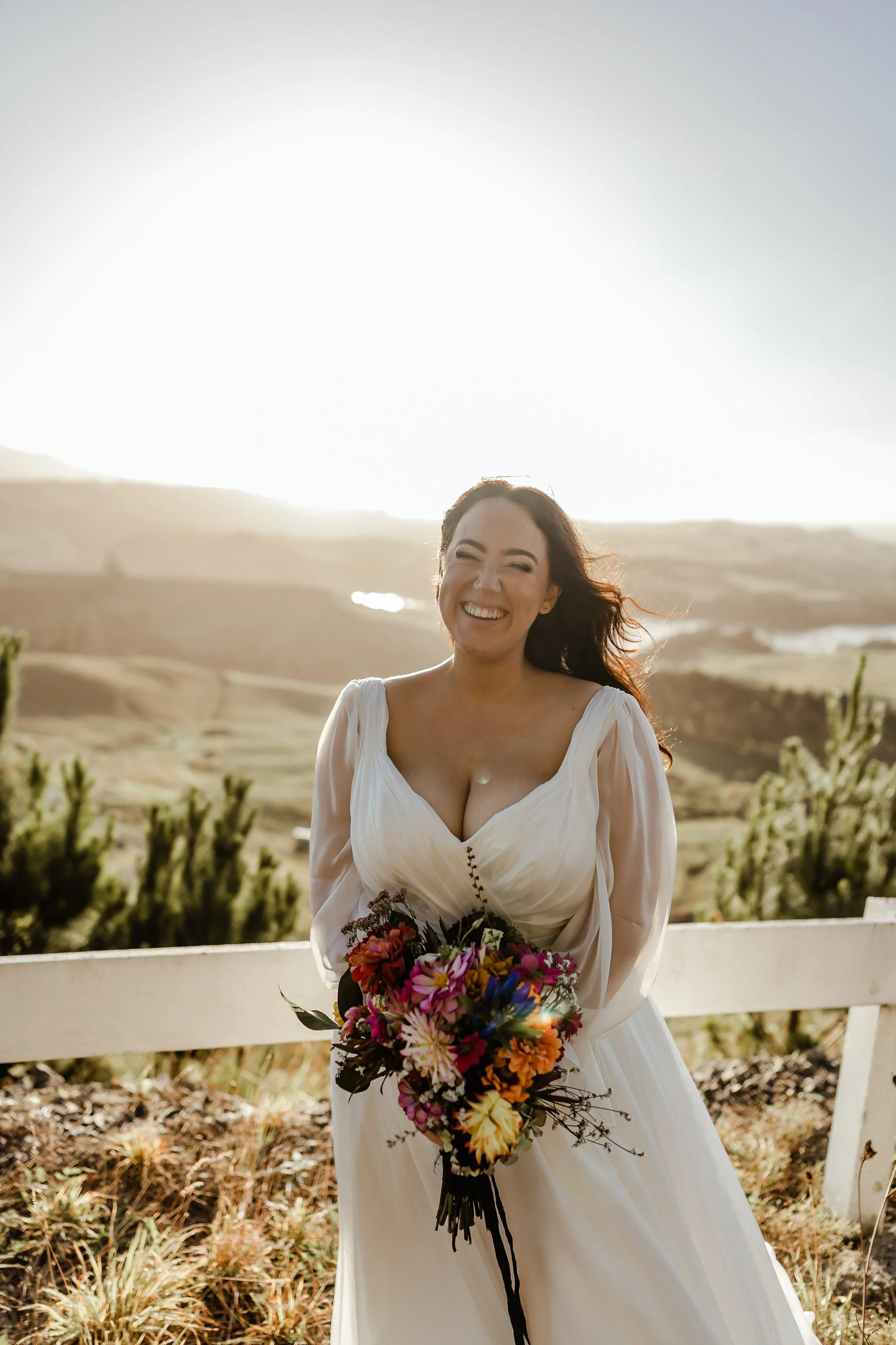 A woman in a white dress holding a colorful bouquet, smiling and standing outdoors with a scenic landscape in the background during sunset.