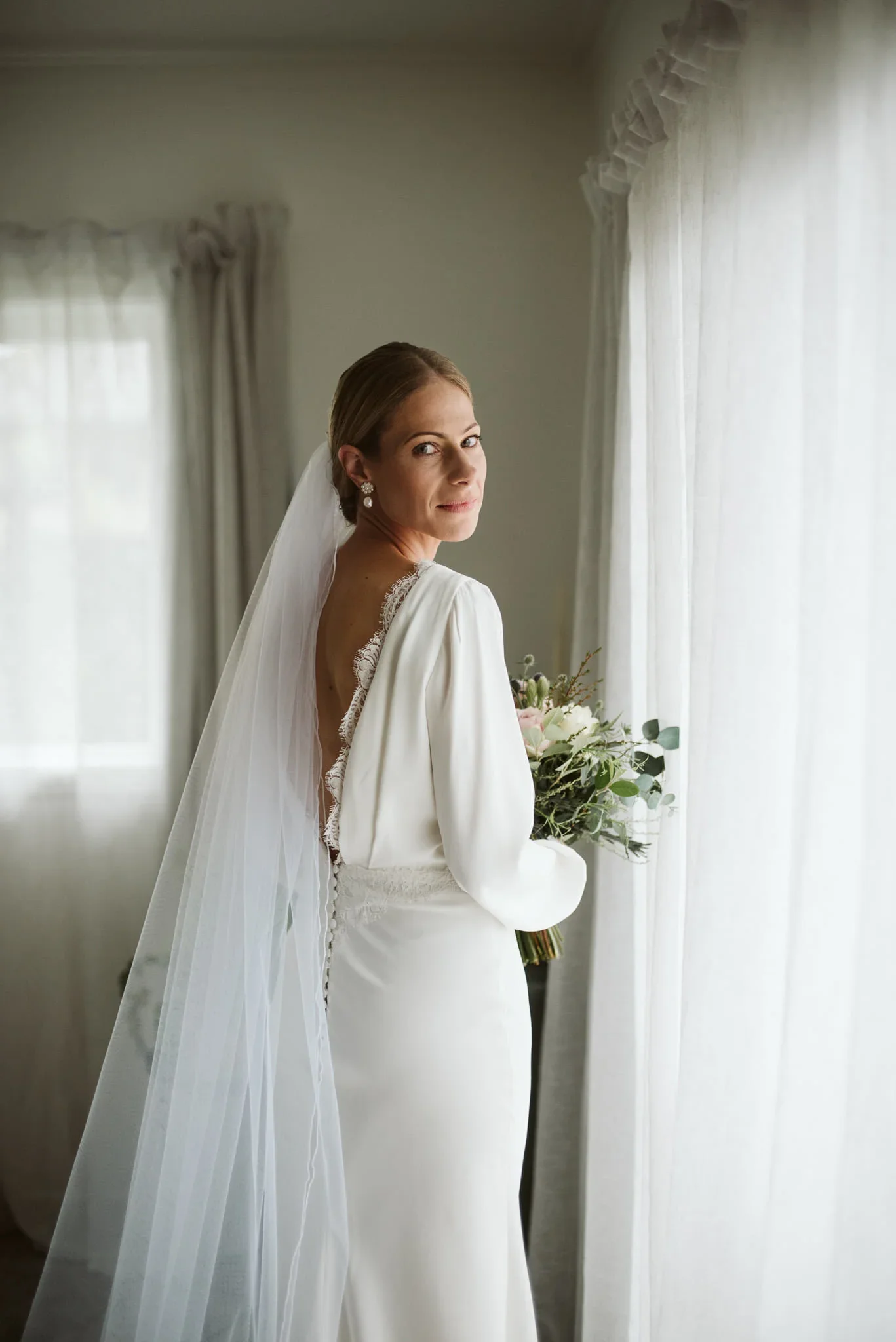 A bride in a white wedding dress with a veil, holding a bouquet of flowers, standing by a window with sheer curtains, looking at the camera.