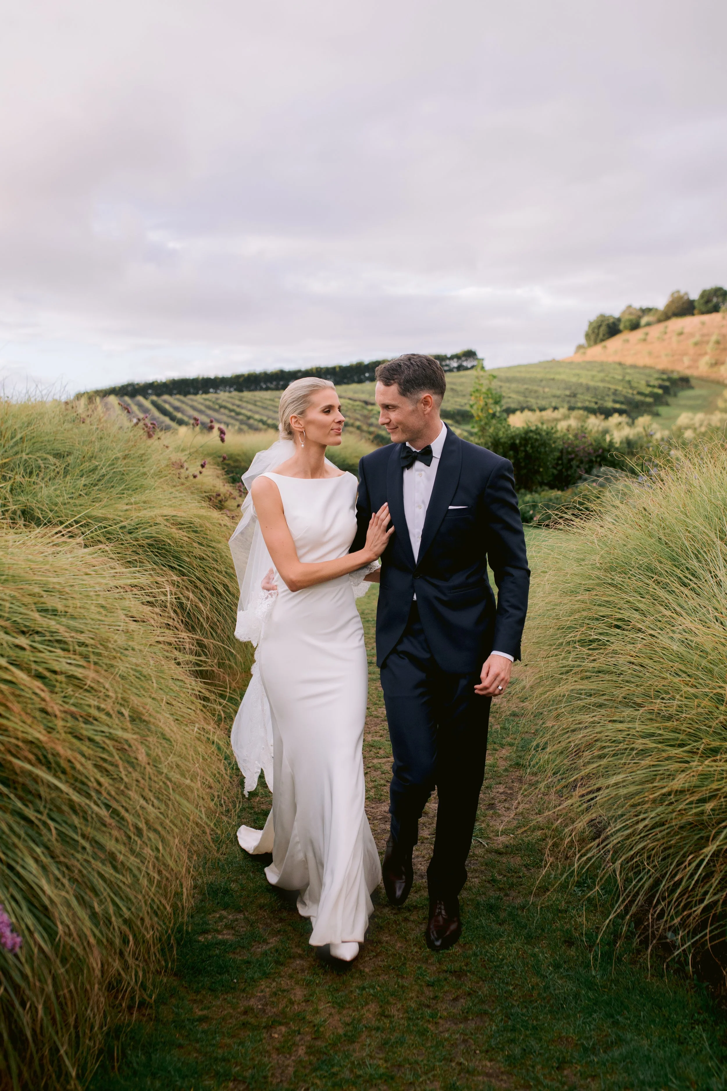 A bride and groom walking together in a field with tall grass and rolling hills in the background.