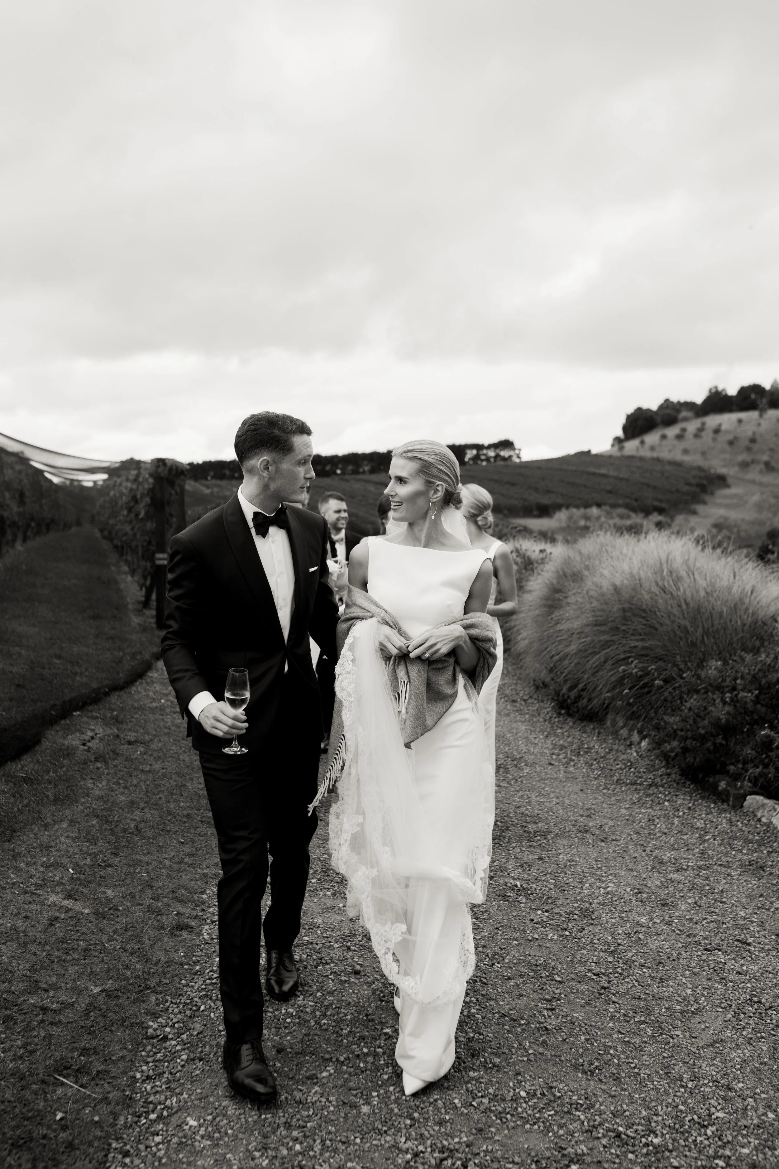 Black and white photo of a bride and groom walking outdoors, the groom in a tuxedo holding a glass of champagne, smiling at each other on a gravel path surrounded by greenery and rolling hills.