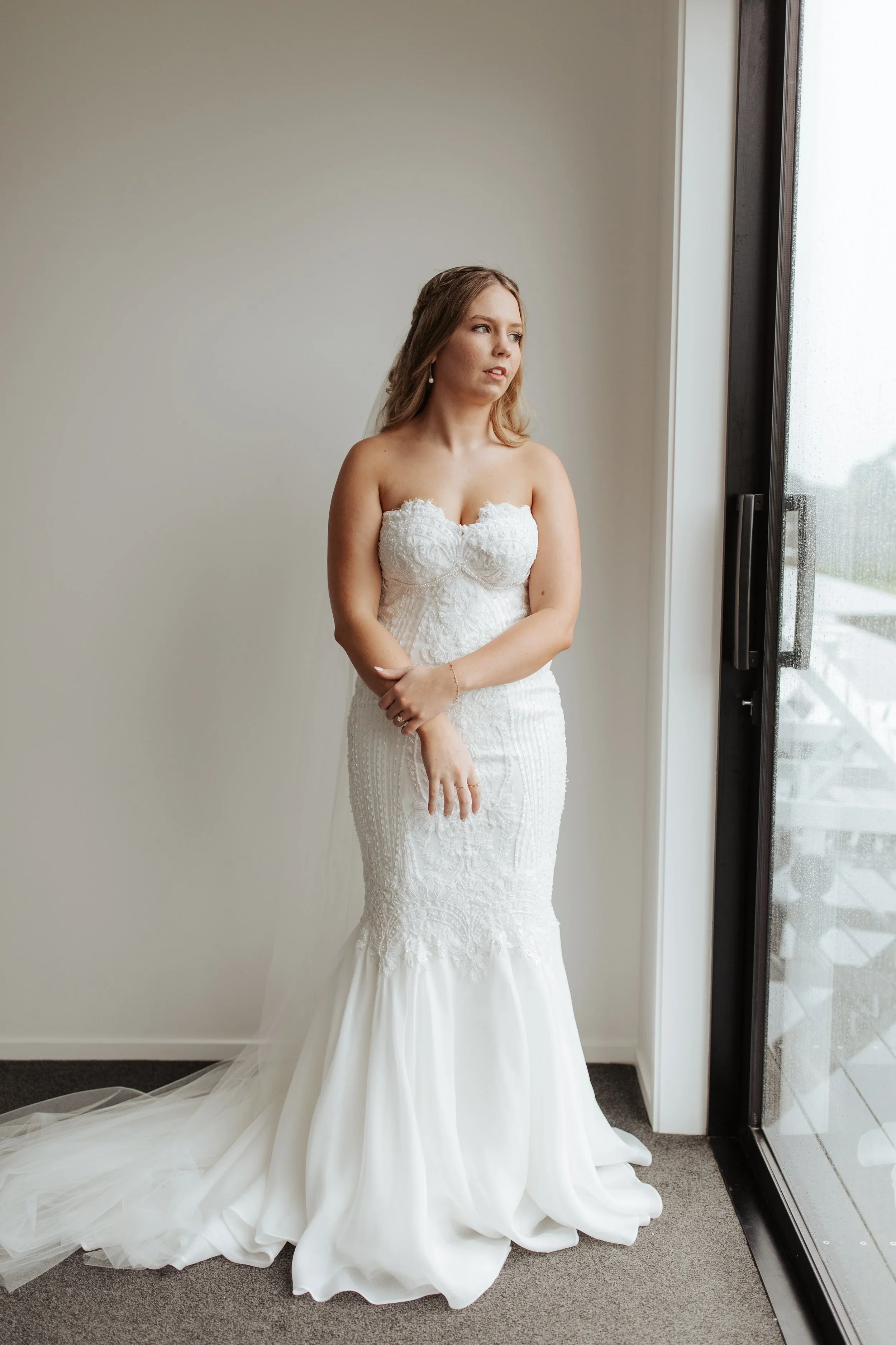 A woman wearing a white wedding dress standing near a floor-to-ceiling window with raindrops on the glass, looking outside.