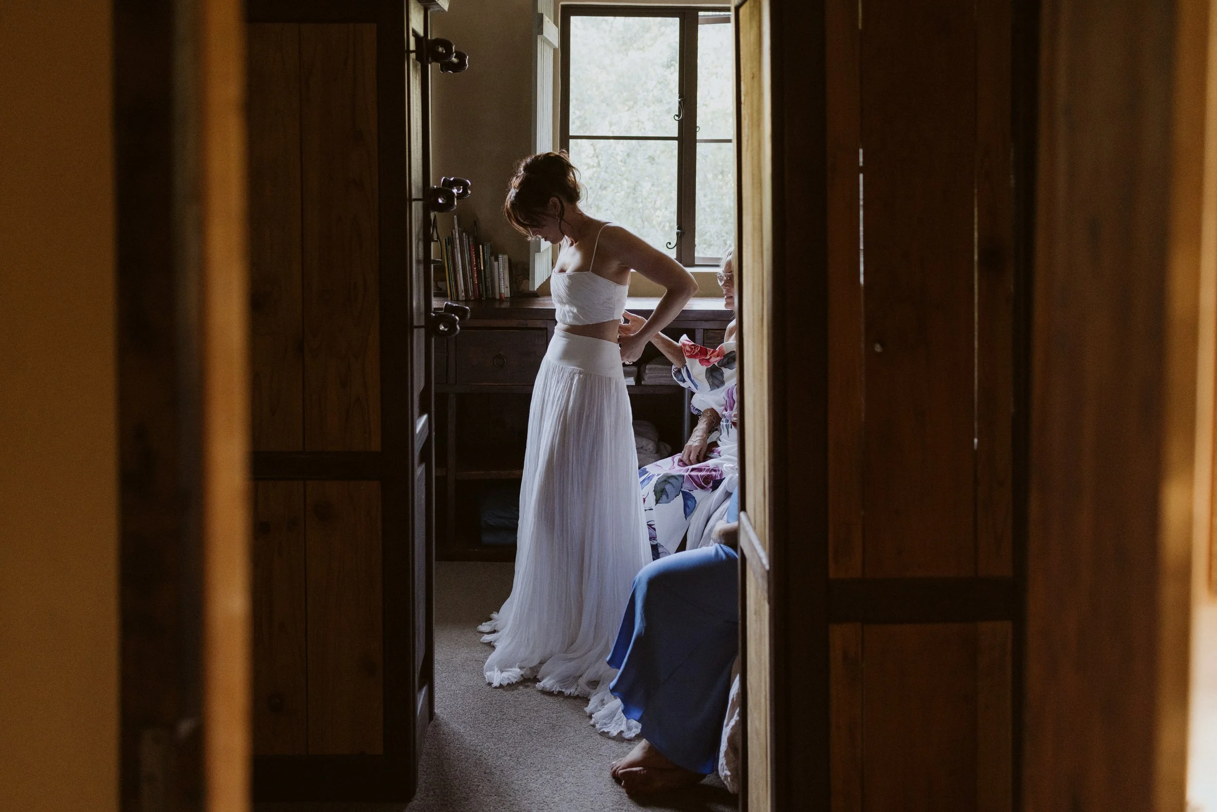 A young woman in a white dress standing in a room with wooden furniture, a window, and books, while an older woman sitting on a chair helps her with the dress.