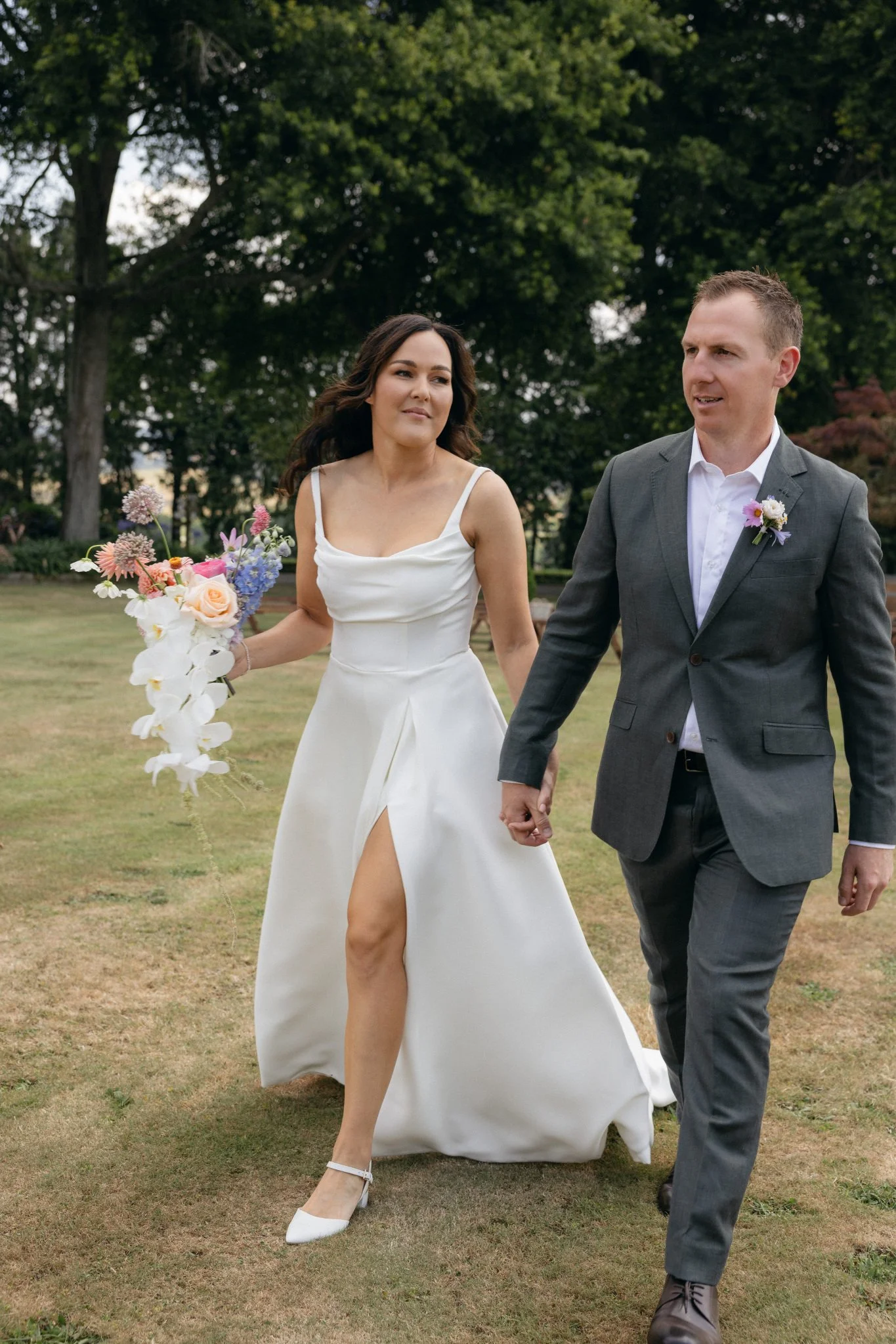 A bride and groom holding hands outdoors, walking on grass in a park with trees, the bride in a white wedding dress with a high slit and the groom in a gray suit with a boutonniere.