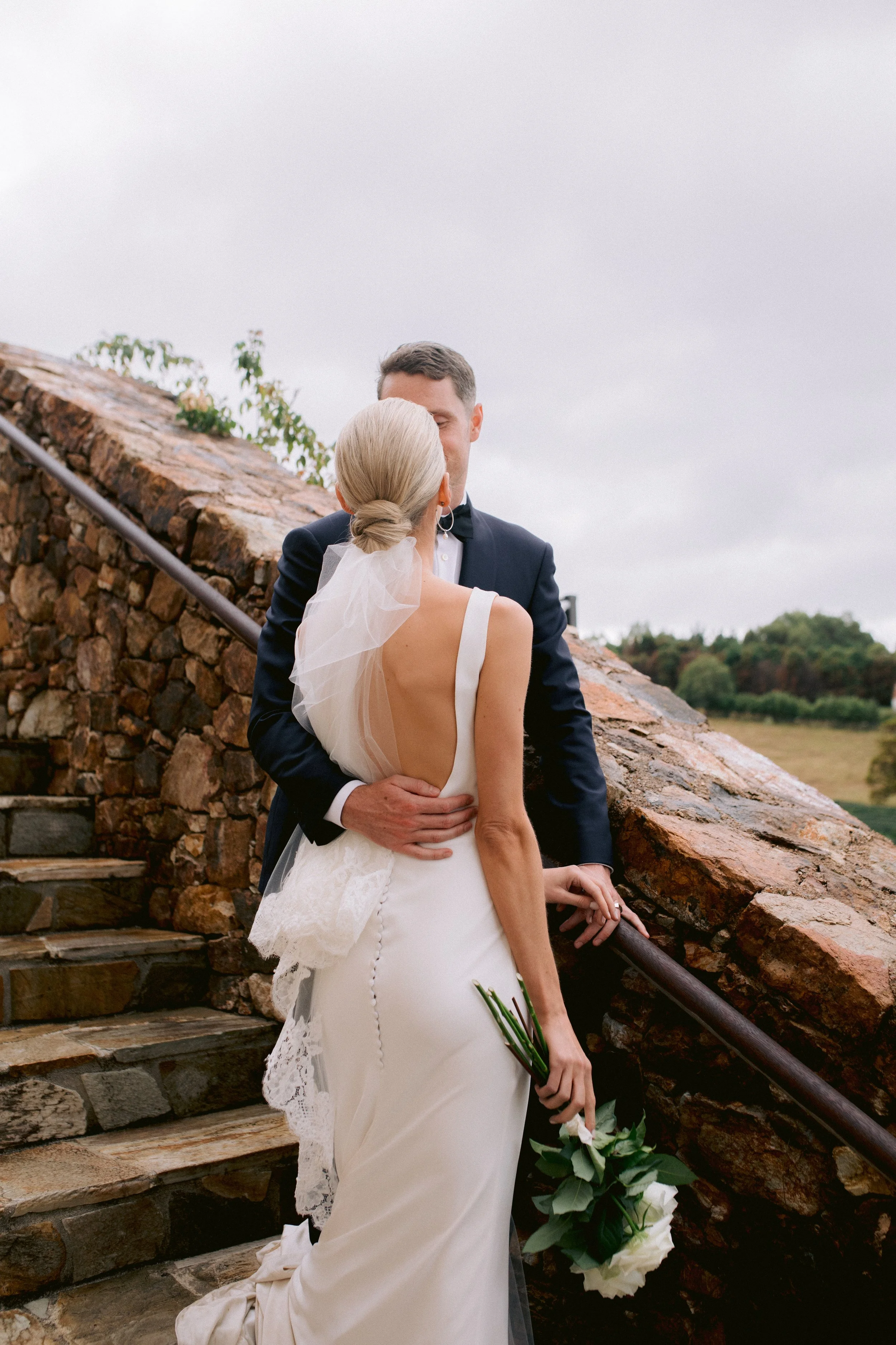 A bride and groom embrace on a stone staircase outdoors during their wedding, with the bride holding a bouquet and wearing a backless white dress with lace details, and the groom in a dark suit.