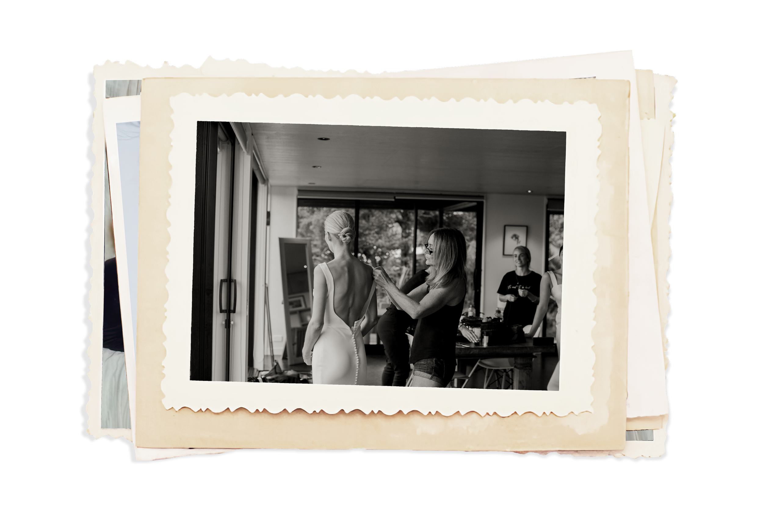 Black-and-white photo of a bride in a wedding dress being assisted by a woman as other women prepare in a room with large windows.